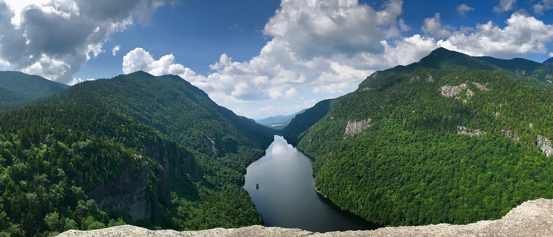 Panoramic view from the Indian Head ledge down Lower Ausable Lake, a long narrow lake enclosed by steep forested mountainsides with cliff bands, under summer cloud
