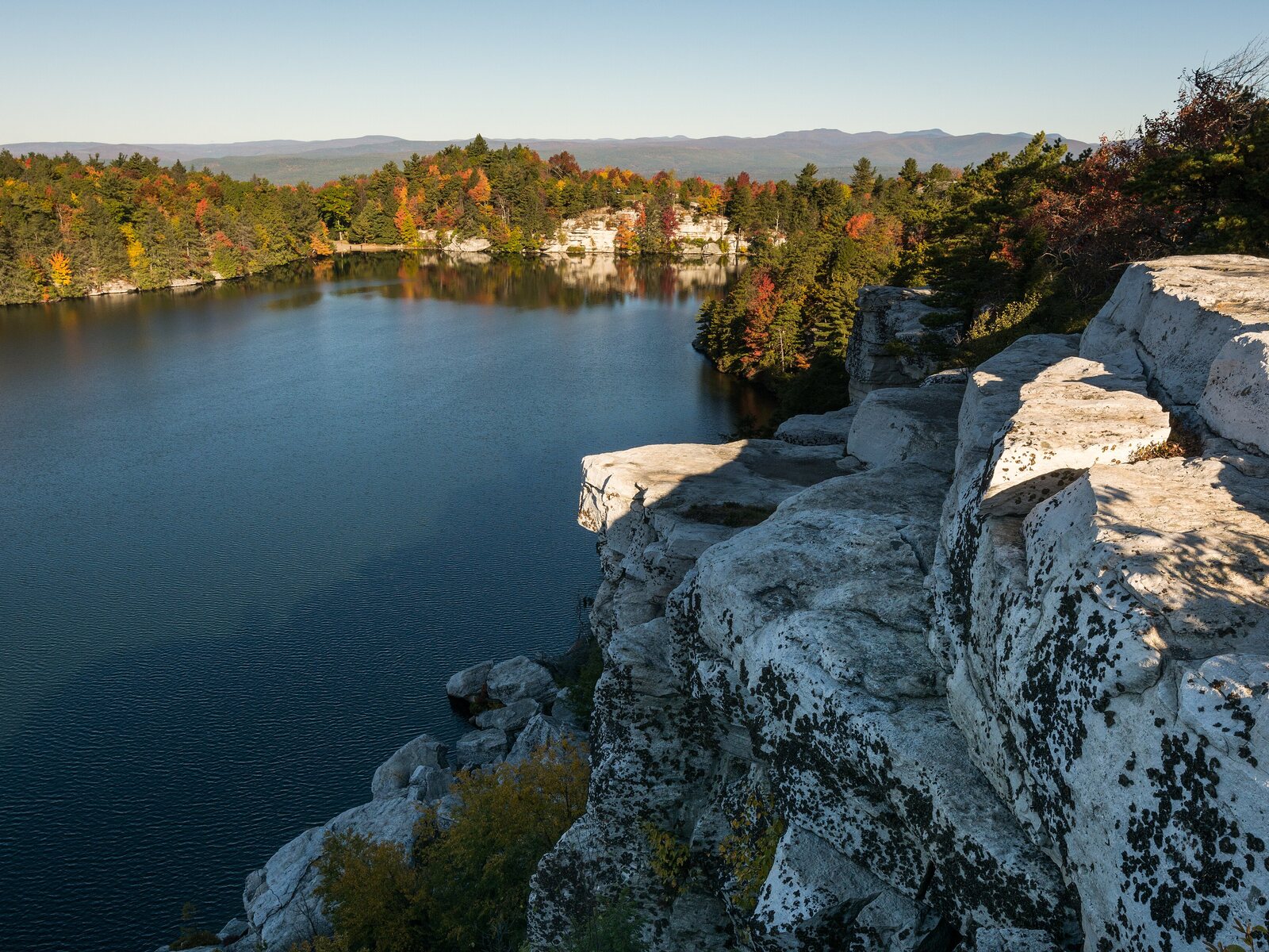 Fall view over Lake Minnewaska from a bare white conglomerate cliff ledge, with orange and green forest around the far shore and the Catskills on the horizon