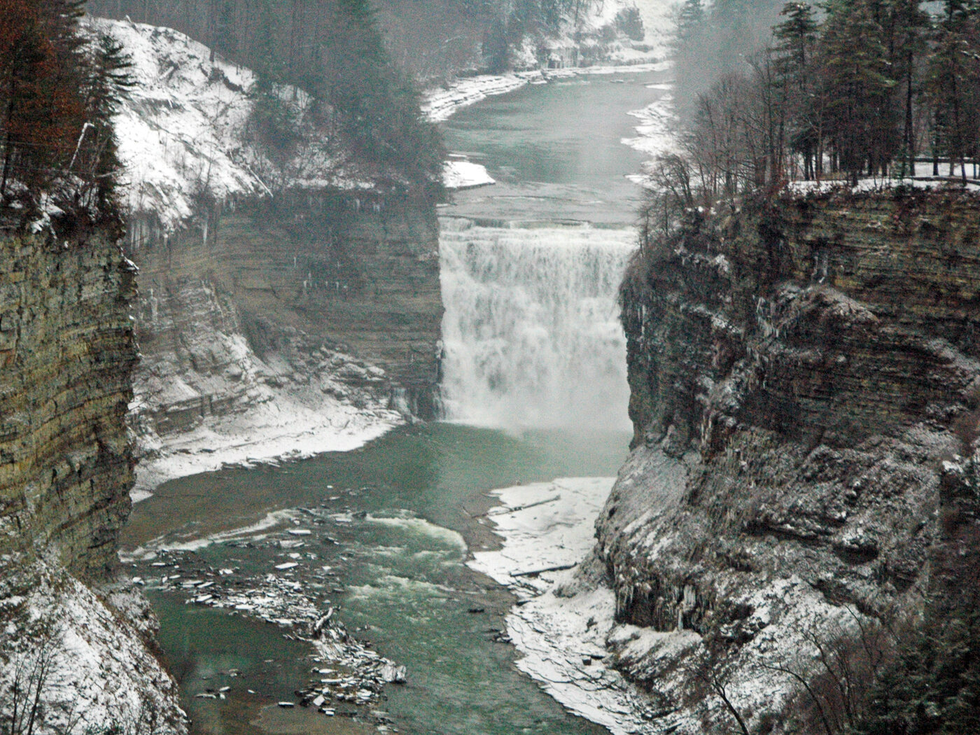 Middle Falls at Letchworth State Park in winter, a wide sheet of water dropping between snow-covered layered cliffs into a green pool