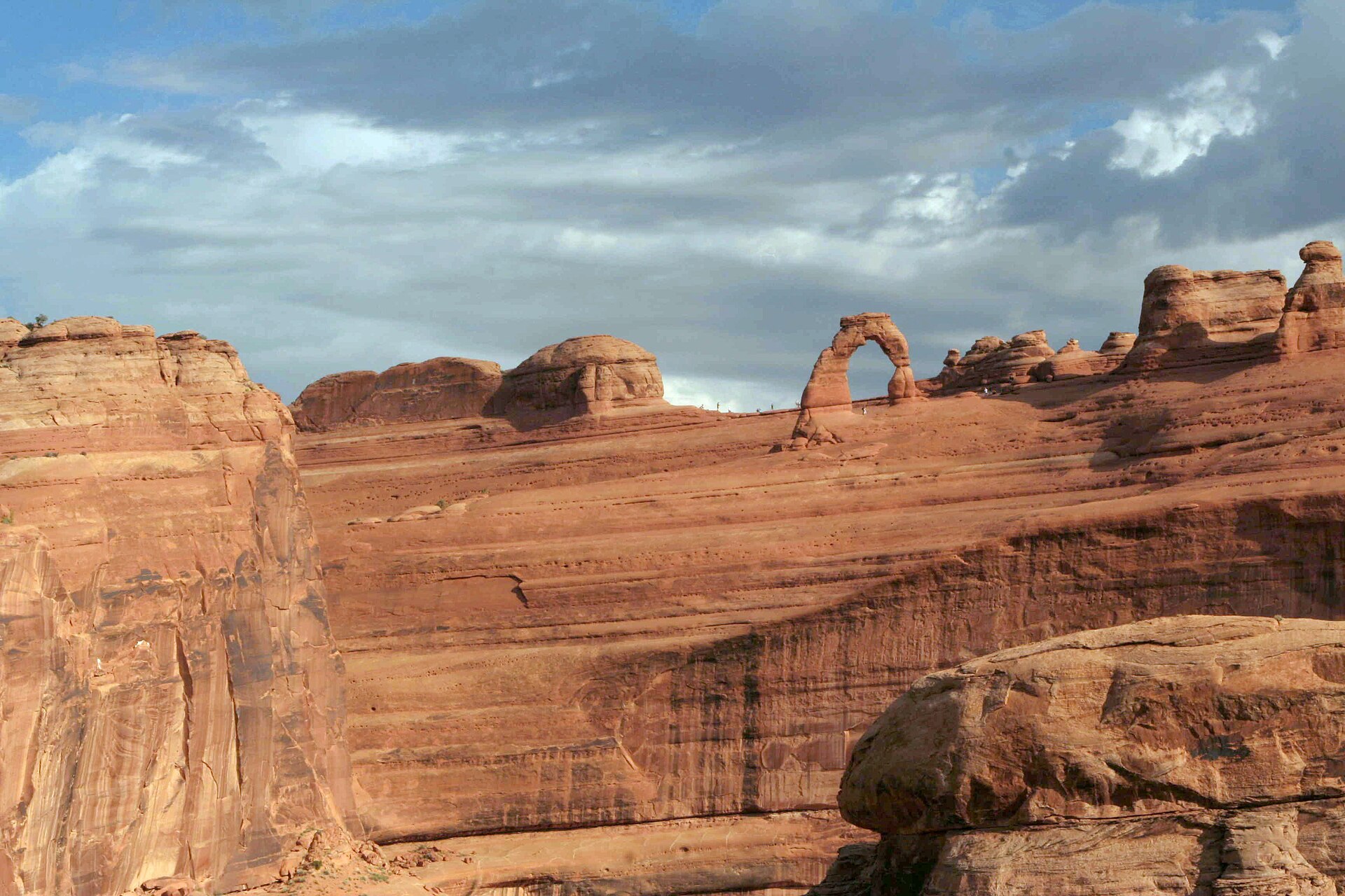 Delicate Arch standing on the rim of a slickrock bowl with tiny figures of hikers nearby, under a bank of cloud
