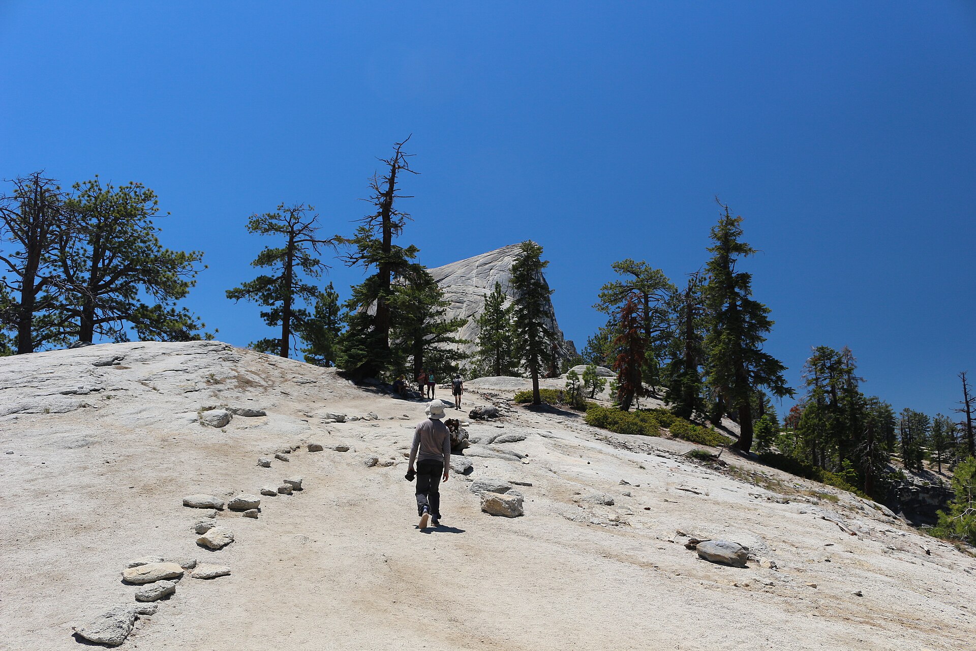 Hikers walking up the bare granite approach toward Half Dome, its rounded summit rising above scattered pines