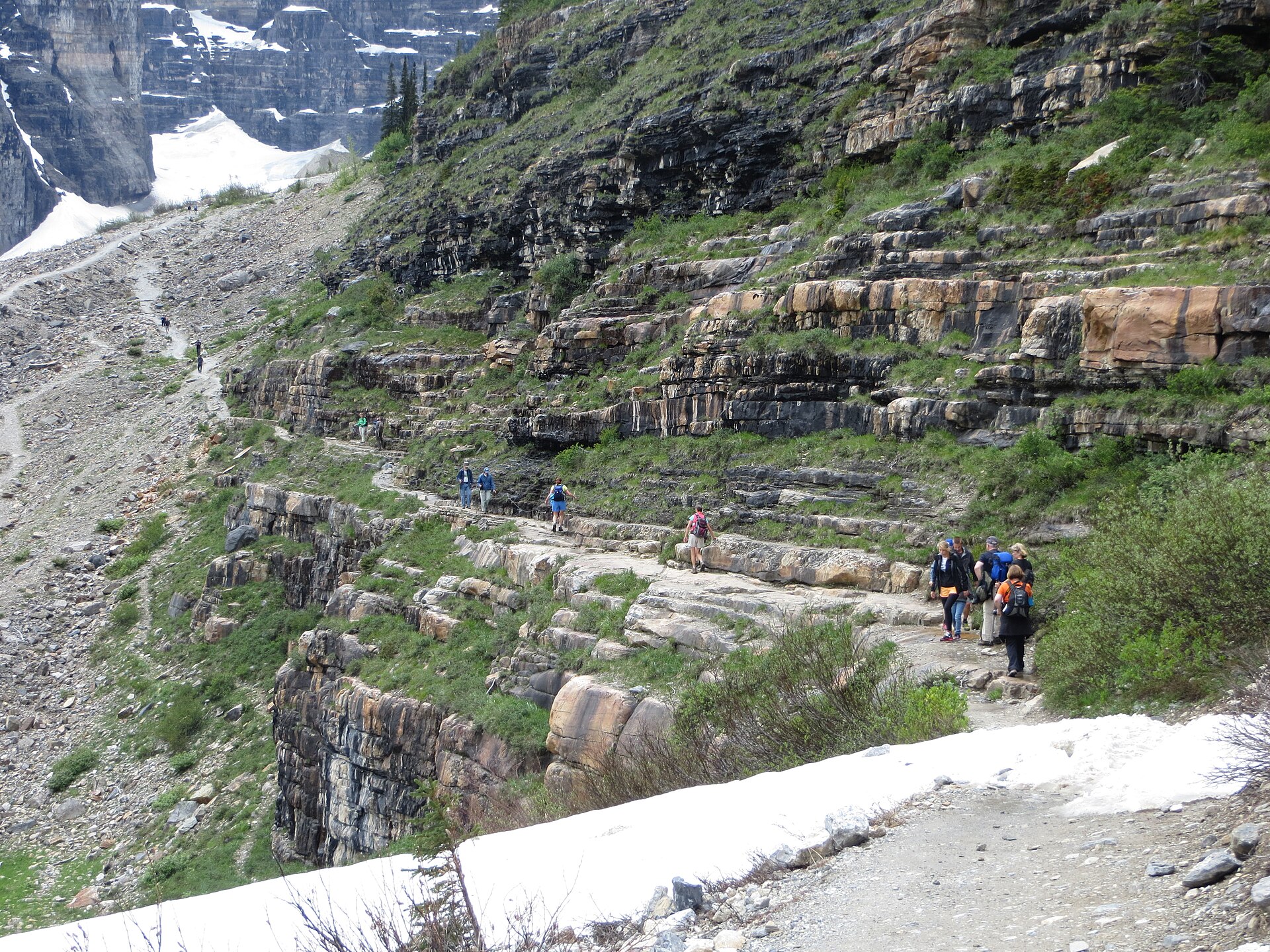 Hikers strung out along the Highline Trail where it is cut into a cliff below the Garden Wall, with snowfields above and below the path