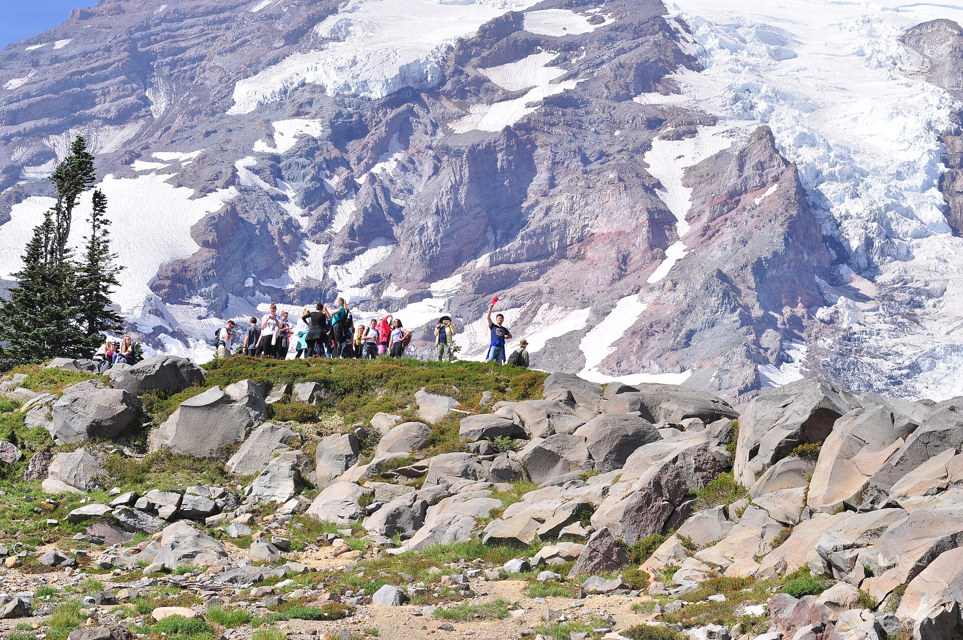 Hikers gathered on a rocky viewpoint on the Skyline Trail with the glaciated bulk of Mount Rainier filling the sky behind them