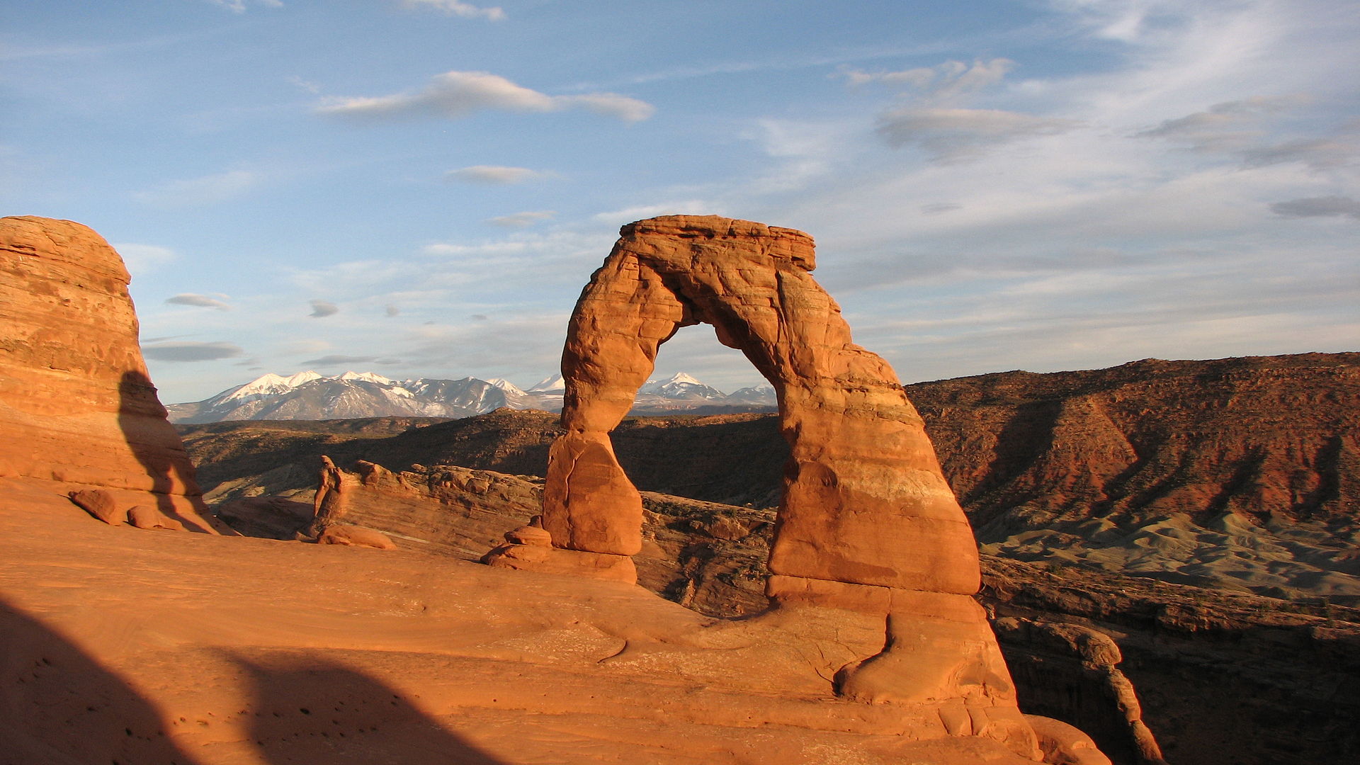 Delicate Arch in Arches National Park at sunset, standing on bare red slickrock with the snow-capped La Sal Mountains behind it