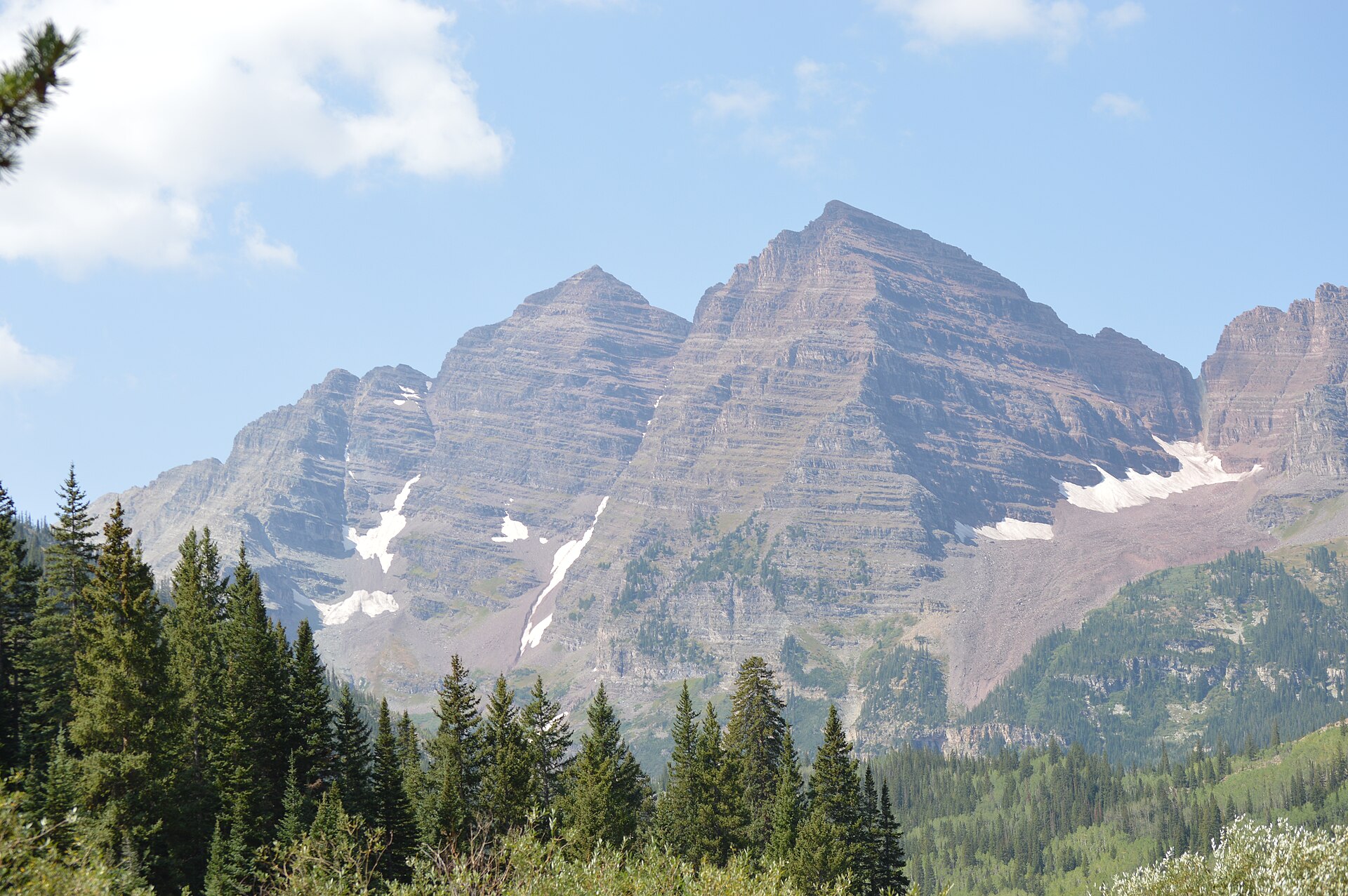 The twin maroon-colored summits of the Maroon Bells above dark spruce forest, streaked with old snow under a bright summer sky