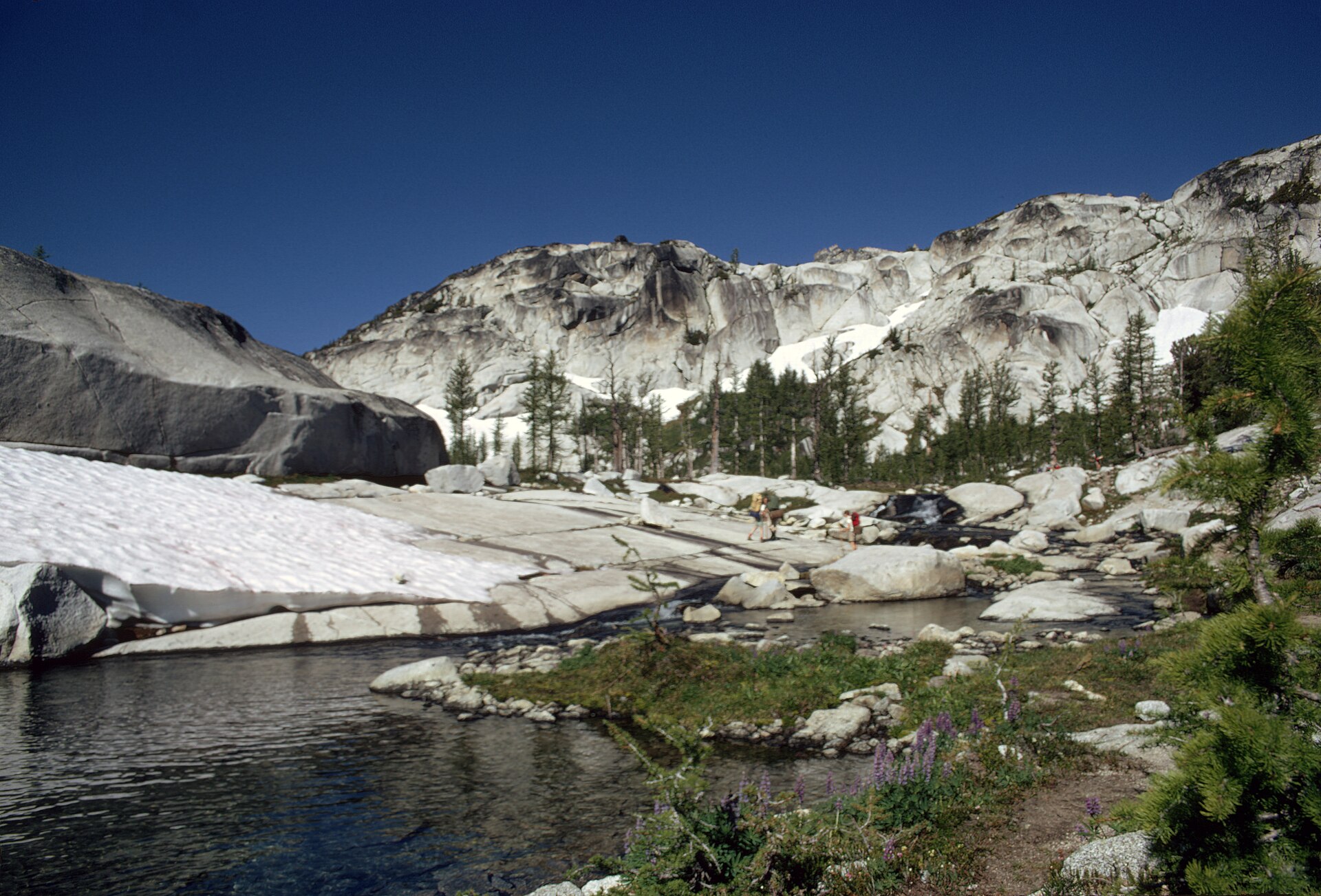 Hikers crossing granite slabs beside an alpine tarn, with old snowfields, scattered larches and a pale granite headwall behind under a deep blue sky