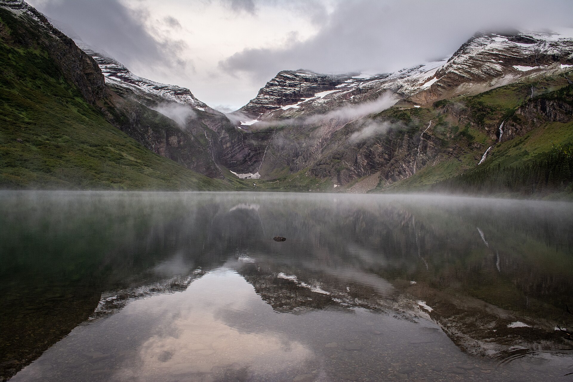 A still alpine lake holding a perfect reflection of a snow-streaked headwall, with mist lying on the water under low grey cloud