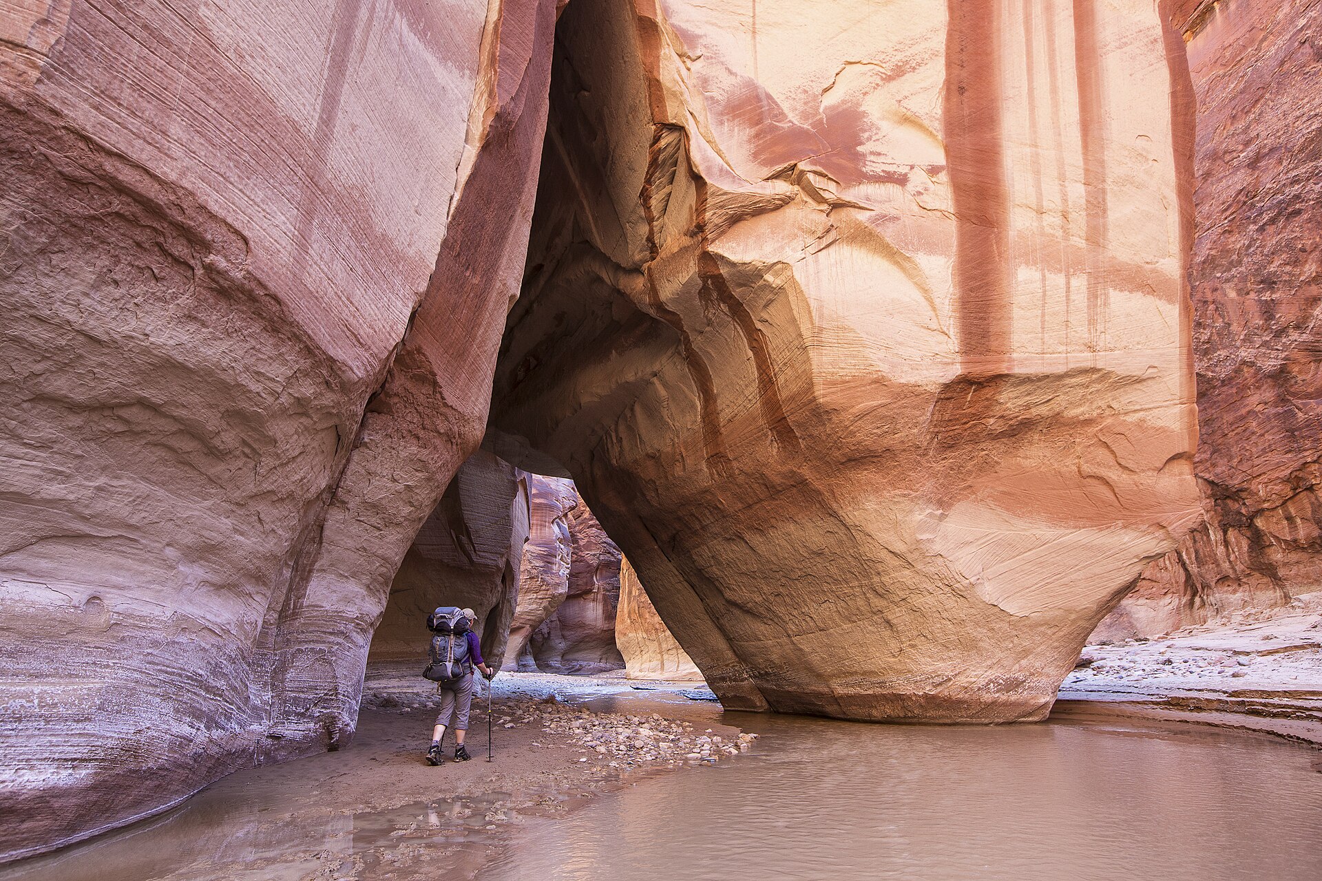 A backpacker standing on the sandy floor of a deep sandstone canyon, dwarfed by curving orange and pink walls that lean together overhead