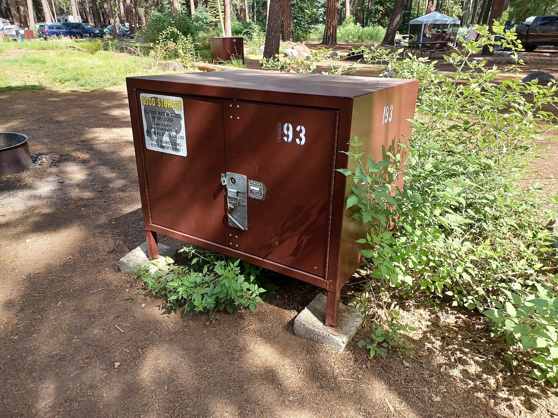 A brown steel bear-resistant food storage locker on concrete footings at a forested campsite in Upper Pines Campground, Yosemite National Park, with a metal latch and a yellow FOOD STORAGE sign on the door