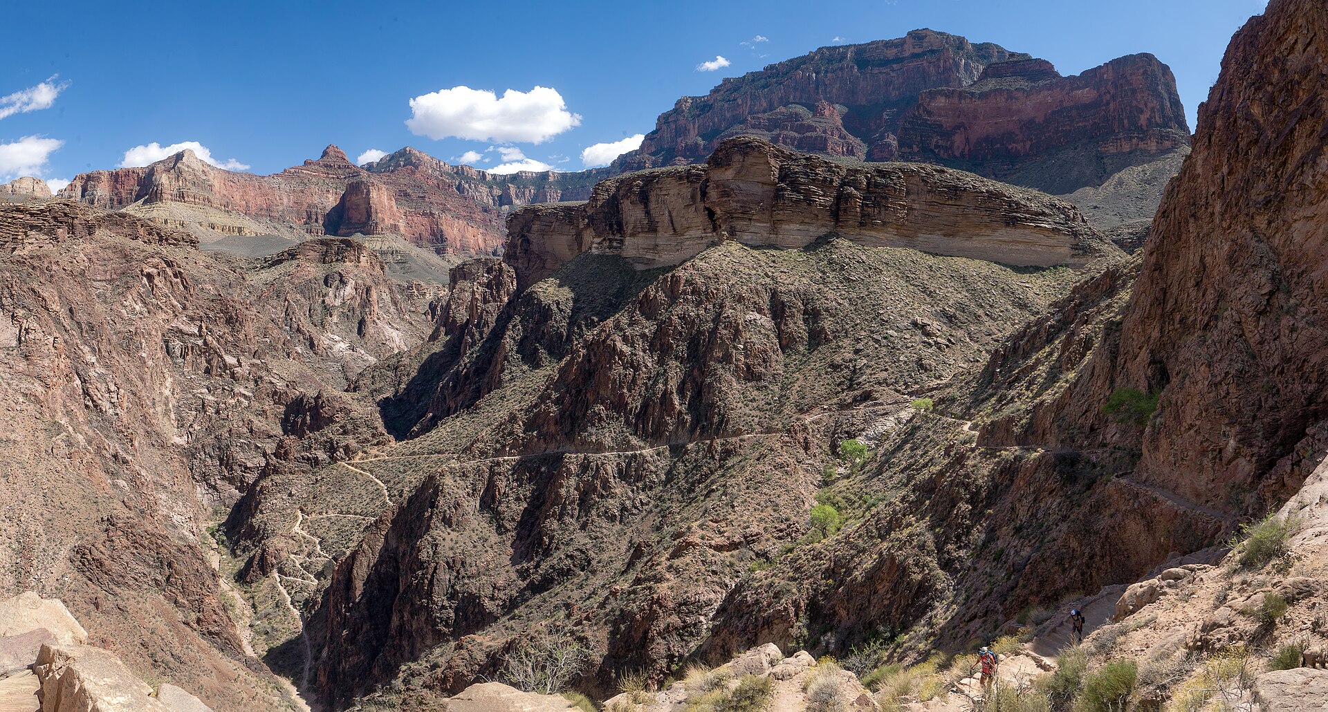 The Bright Angel Trail in Grand Canyon National Park threading in pale switchbacks across bare rock and scrub above Pipe Creek, with two tiny hikers on the trail at lower right and no shade anywhere in the frame