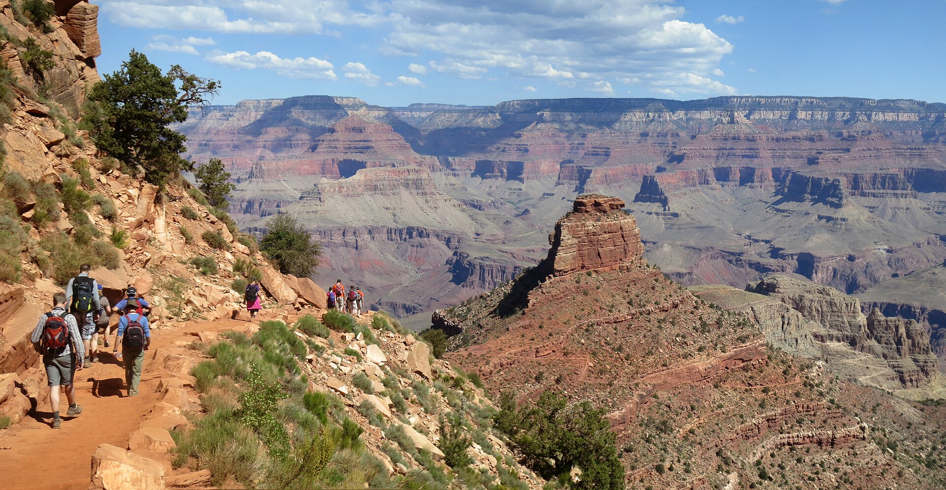 A line of day hikers descending the South Kaibab Trail in Grand Canyon National Park on open, unshaded rock, with O'Neill Butte and the far canyon walls behind them