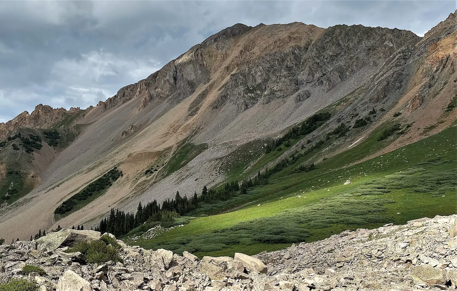 The southwest face of Mount Daly in the Maroon Bells-Snowmass Wilderness, a long scree slope under a rocky summit with green meadow and scattered conifers below