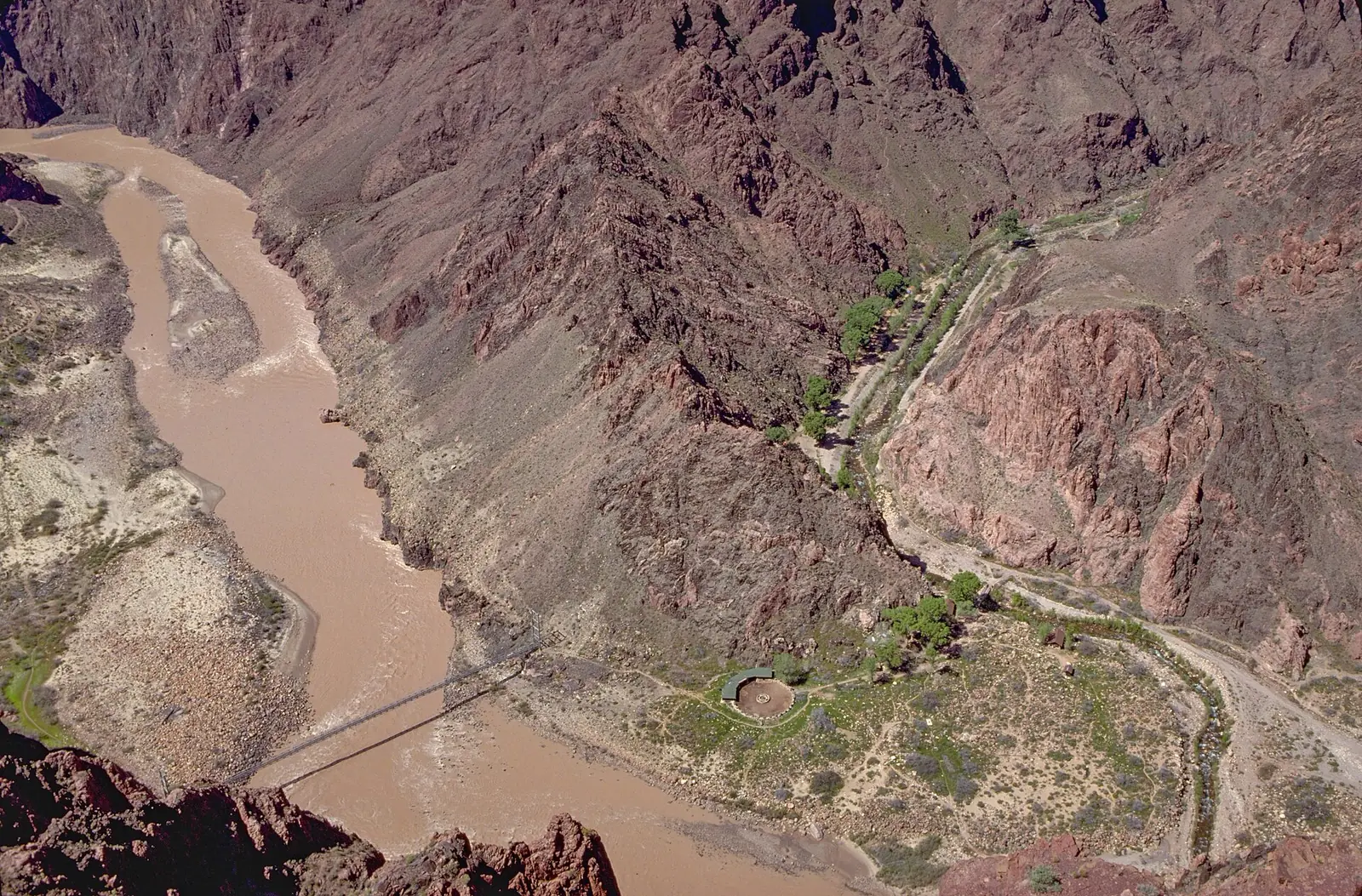 Aerial view of the Silver Bridge crossing the muddy Colorado River, with the green cottonwoods of Bright Angel Campground along the creek on the far bank