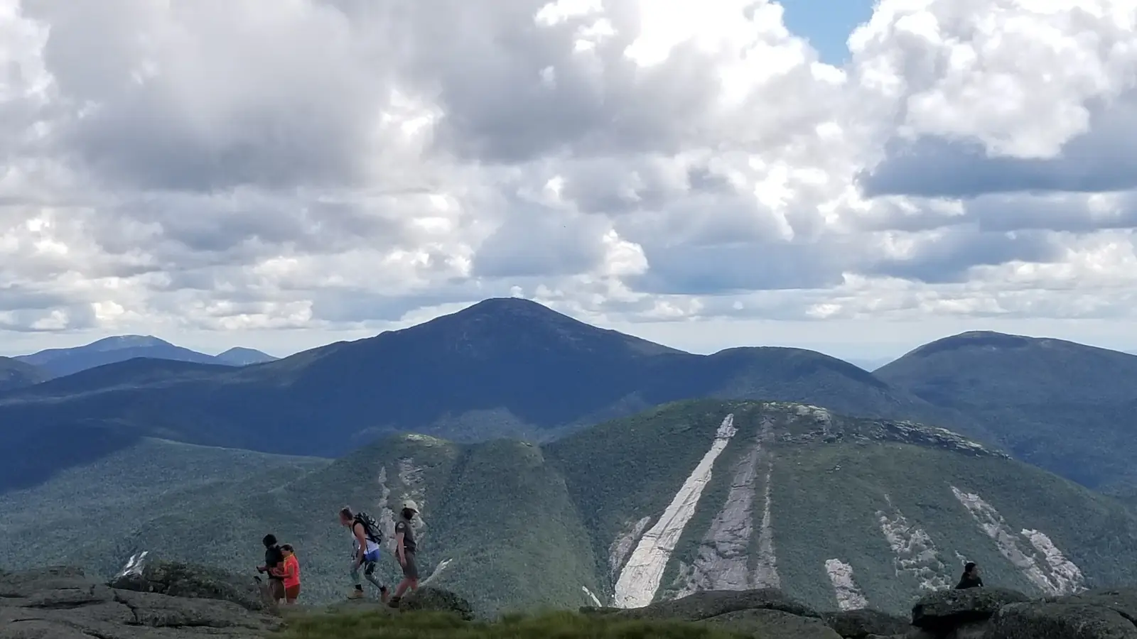 Hikers walking across the bare rock summit of Algonquin Peak, with Mount Marcy behind and the white slides of Mount Colden falling away to the right