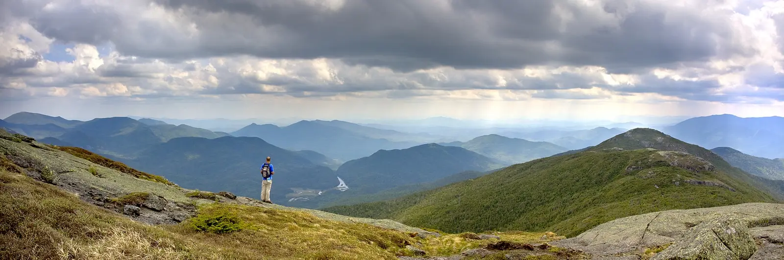 A single hiker standing on the open alpine summit of Algonquin Peak, looking out over ridge after ridge of forested Adirondack mountains under a heavy sky