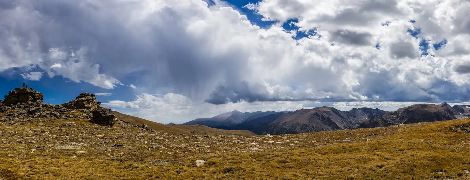 Golden alpine tundra above the treeline in Rocky Mountain National Park, with a rocky outcrop on the left, a line of distant peaks and a storm building over the divide