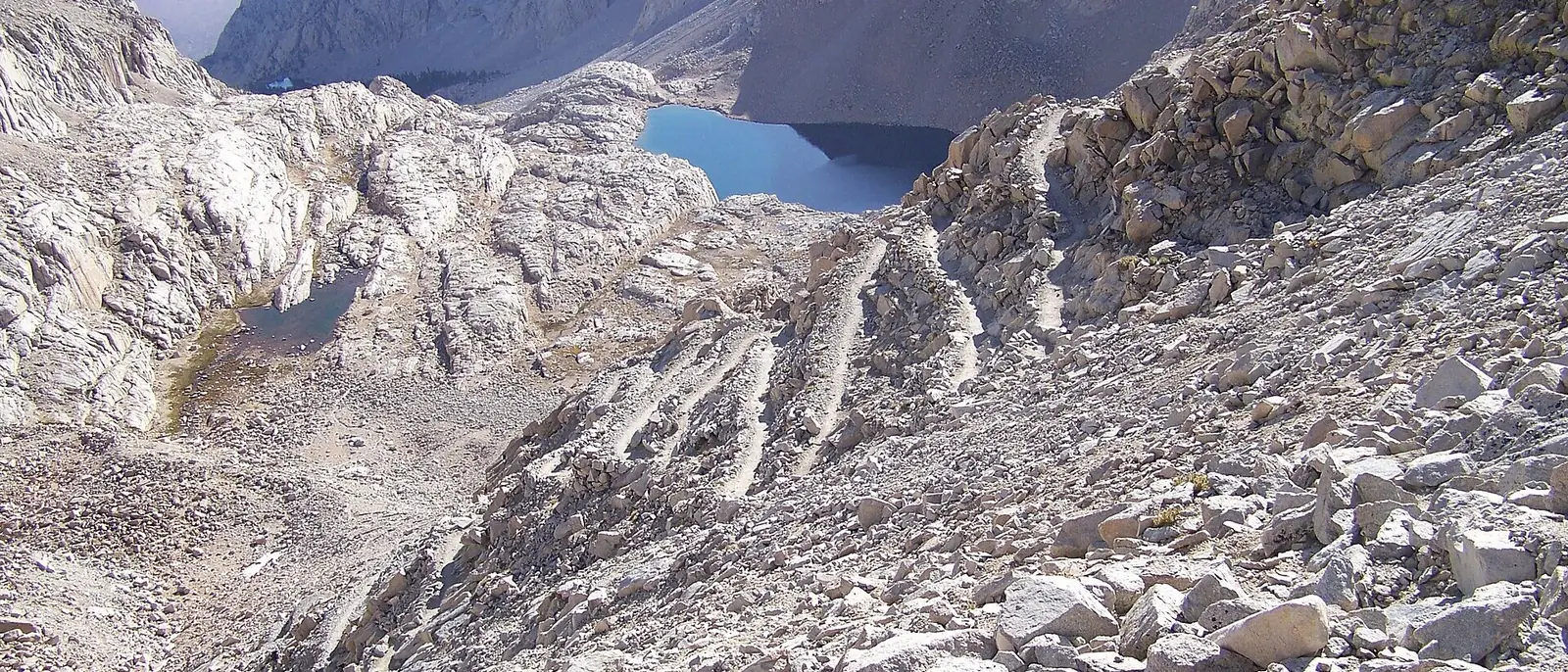 Switchbacks cut into pale granite on the Mount Whitney Trail, climbing away from a blue alpine lake in a bare rocky basin