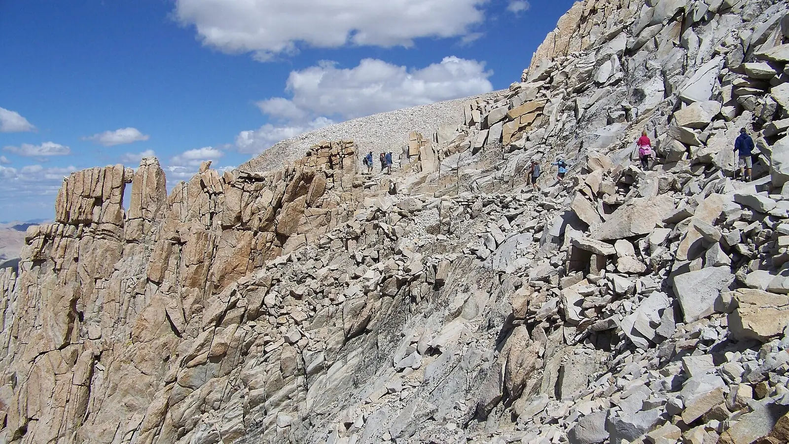 Hikers strung out across bare pale granite on the final traverse of the Mount Whitney Trail, with the fractured rock windows of the summit ridge on the left
