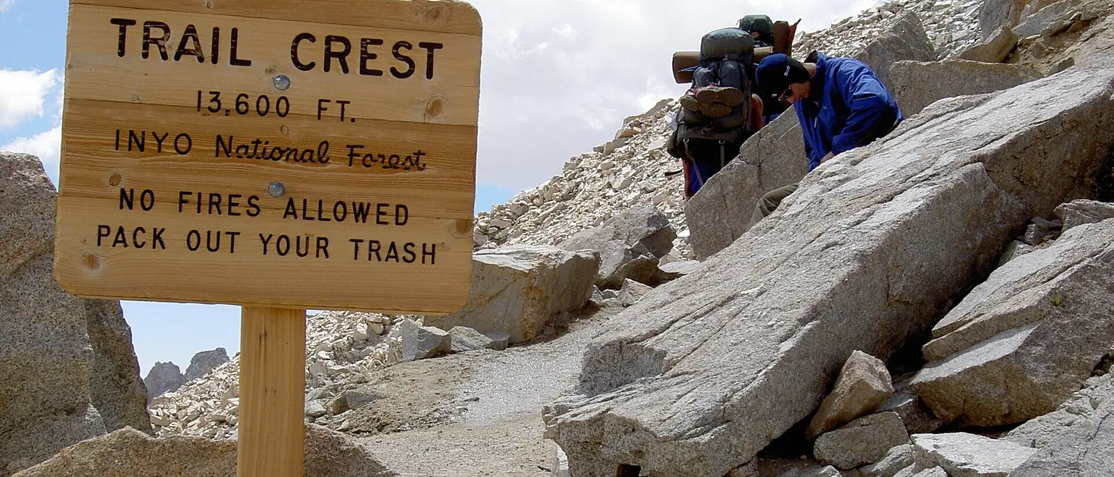 A wooden Forest Service sign reading Trail Crest 13,600 ft beside a sandy trail, with two loaded backpackers picking their way across granite blocks behind it