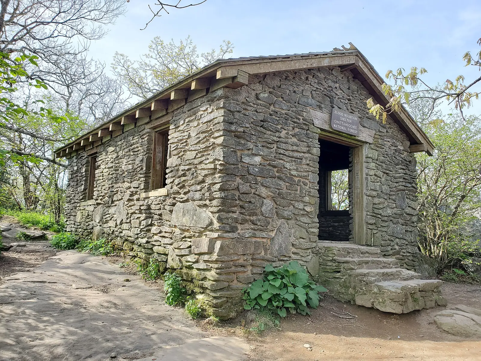 A low stone shelter with a shingled roof and open doorway on the wooded summit of Blood Mountain, Georgia, with a carved sign reading Blood Mountain Shelter Chattahoochee National Forest