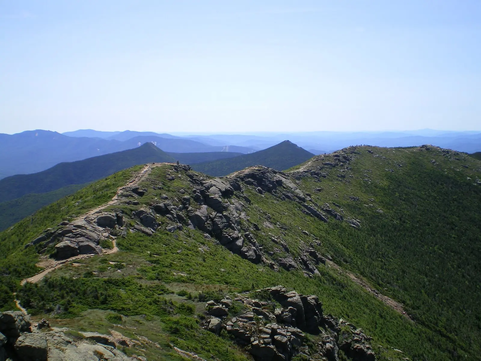 The Appalachian Trail running along the bare rocky crest of Franconia Ridge in New Hampshire above the treeline, green krummholz on the slopes and blue mountain ranges receding to the horizon