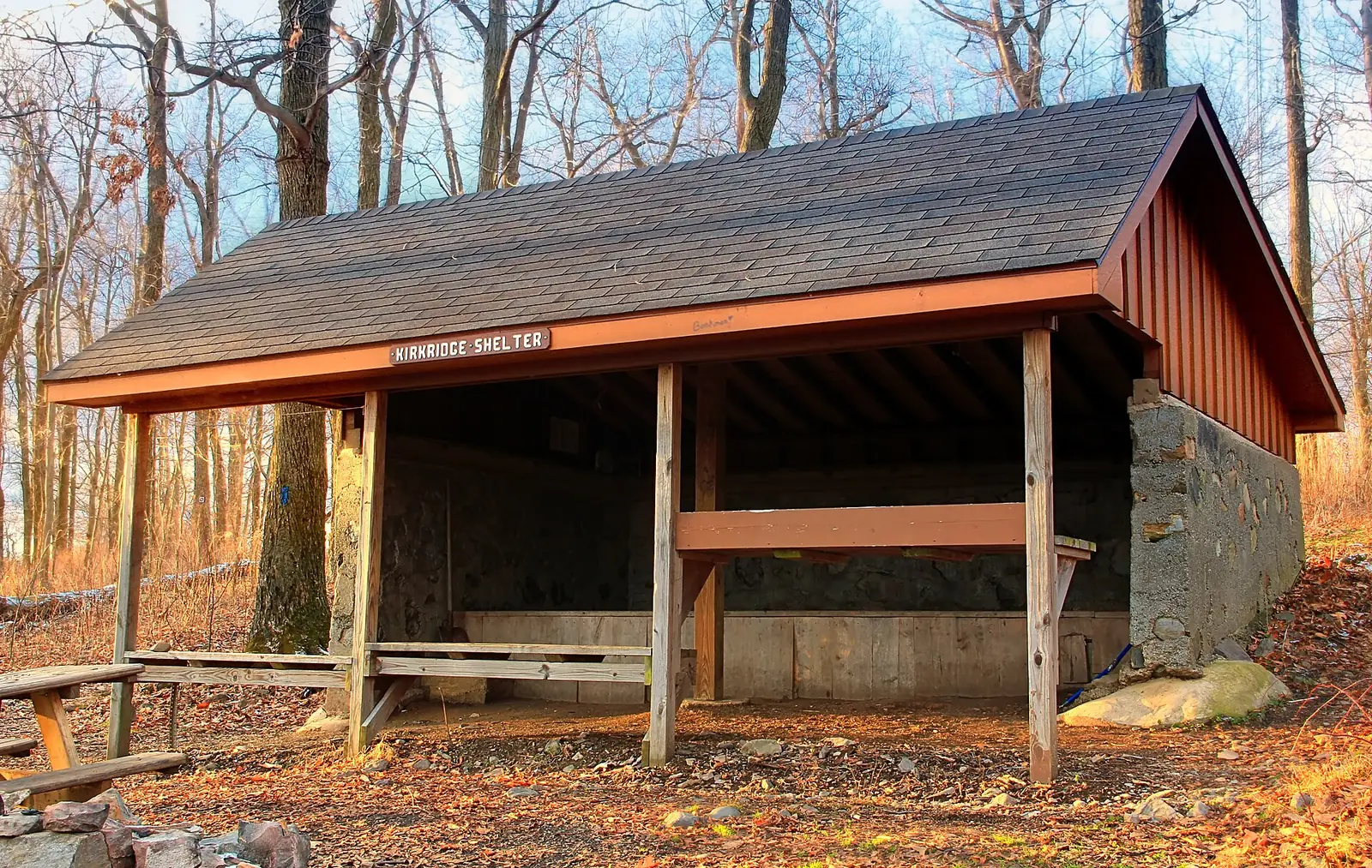 A three-sided wooden shelter with a shingled roof and a sign reading Kirkridge Shelter, open along its long side to show a raised sleeping platform and a bench, with bare winter trees behind