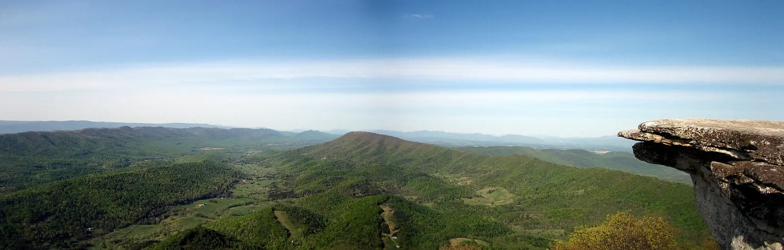 The wide view west from McAfee Knob in Virginia, green ridges and farm valleys under a pale sky, with the flat overhanging ledge of the knob jutting into frame on the right