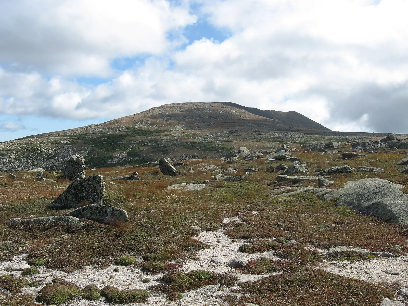 The bare granite dome of Baxter Peak on Katahdin seen across the open alpine tableland, boulders and low tundra vegetation in the foreground under a bright cloudy sky