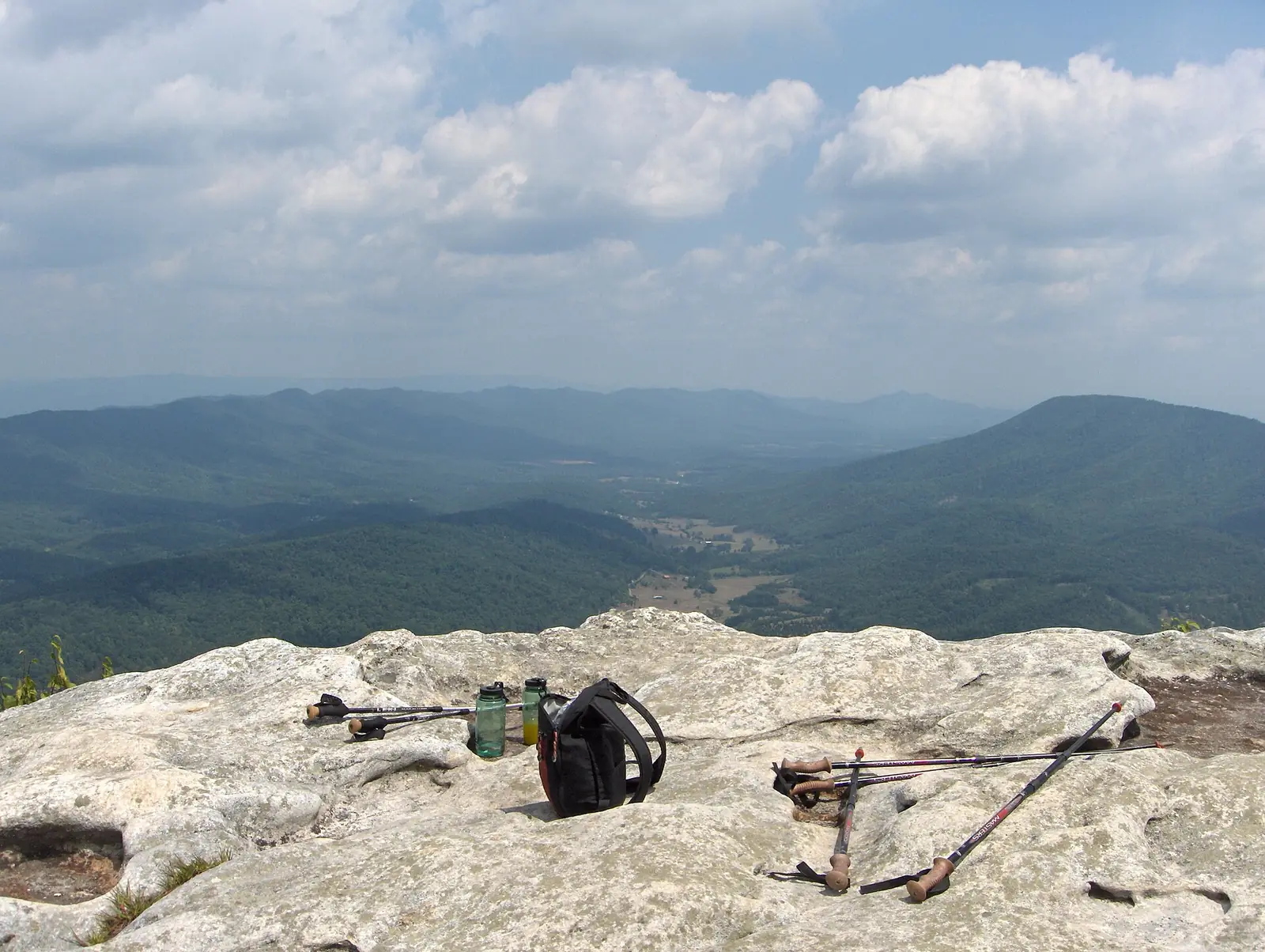 The broad flat summit rock of McAfee Knob in Virginia with a daypack, two water bottles and two pairs of trekking poles resting on it, looking out over the green Catawba Valley and blue ridges under cloud
