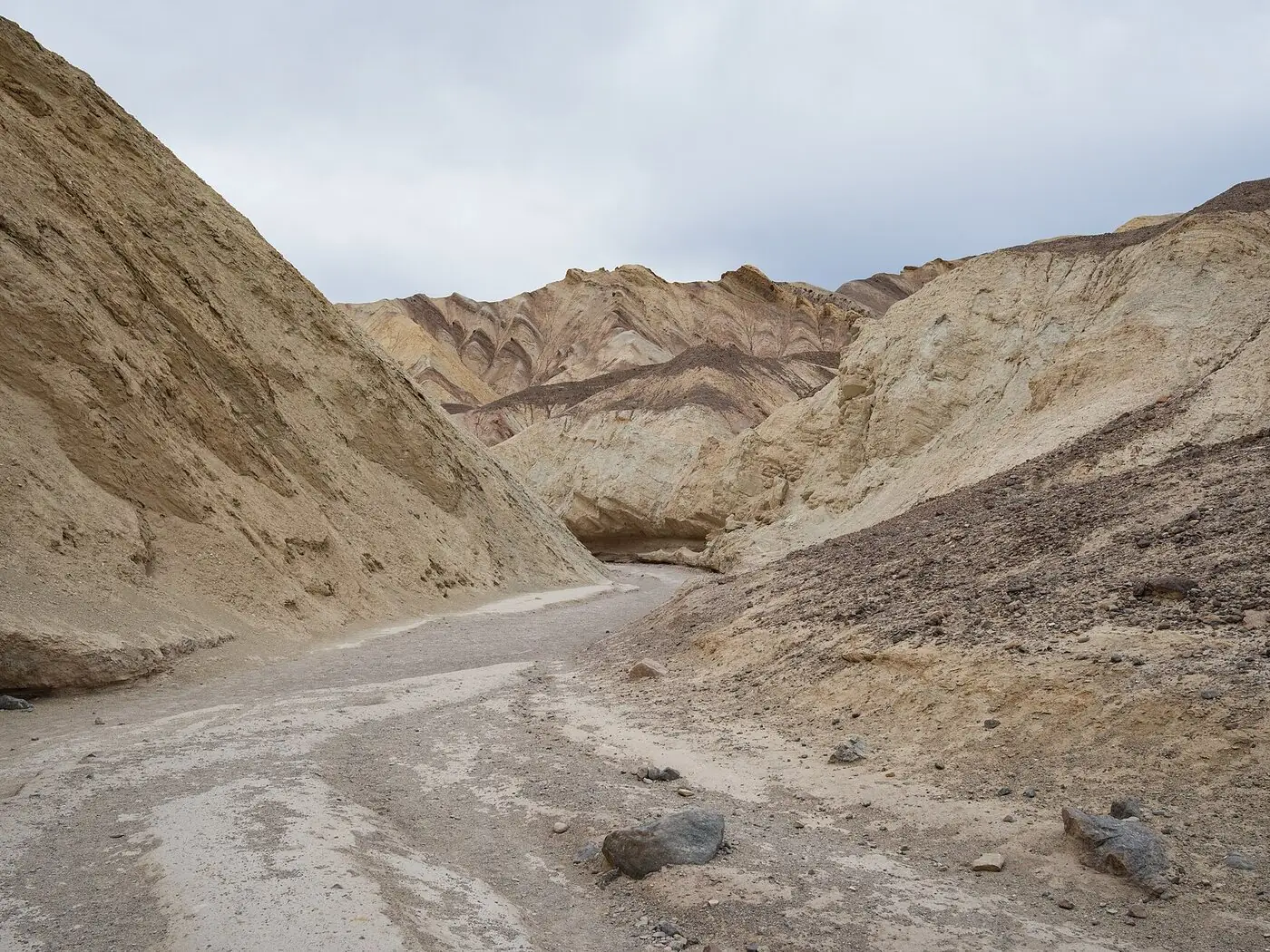 A dry gravel wash running between pale eroded badland walls in Golden Canyon, Death Valley, with banded ridges of cream, purple and brown rock behind