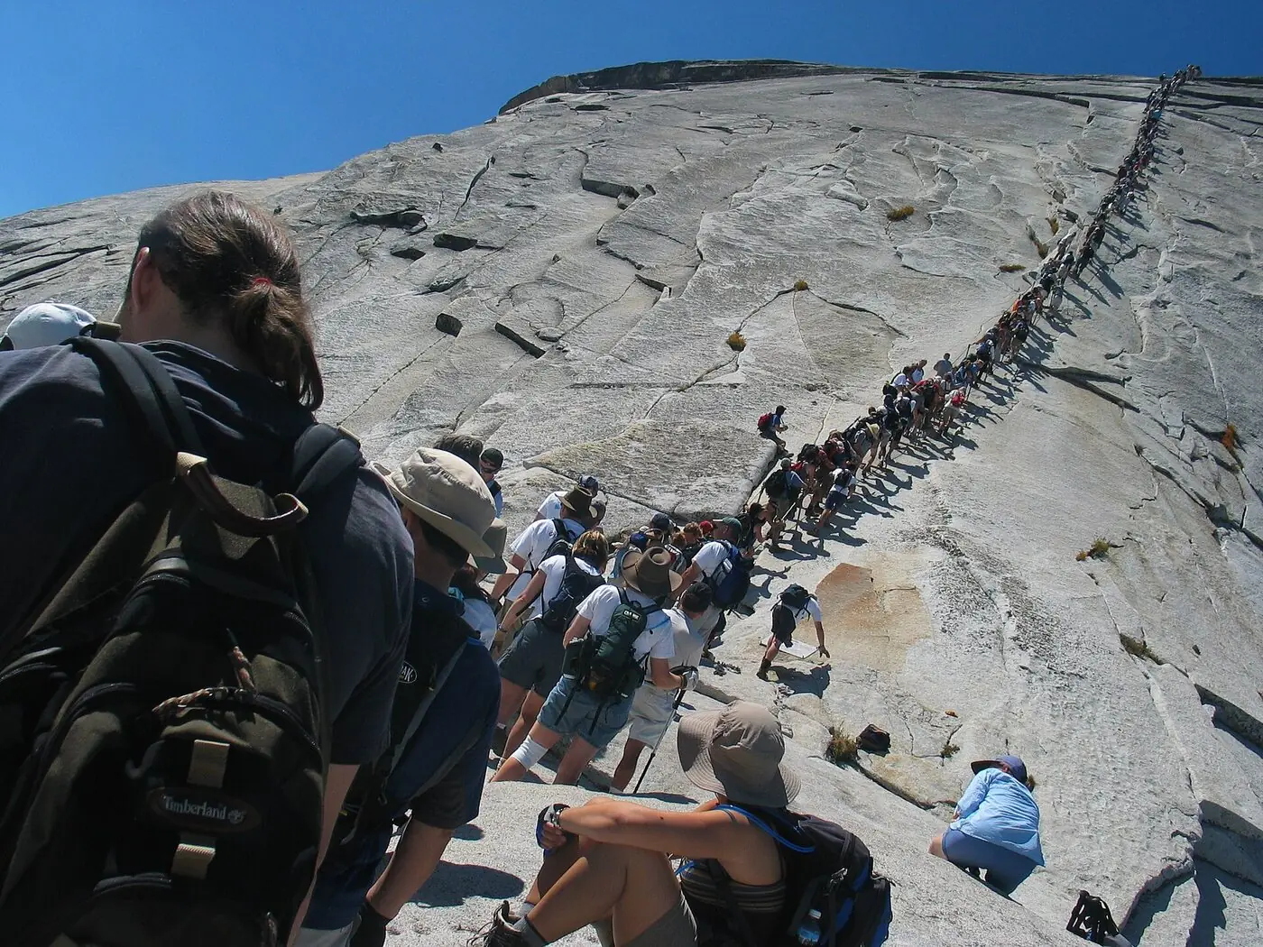 A long queue of hikers strung out along the two steel cables running up the steep granite flank of Half Dome, with more hikers waiting on the rock below