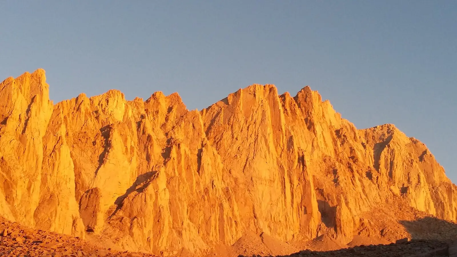 The east face of Mount Whitney lit orange by sunrise, seen from Trail Camp on the Mount Whitney Trail against a clear pale sky