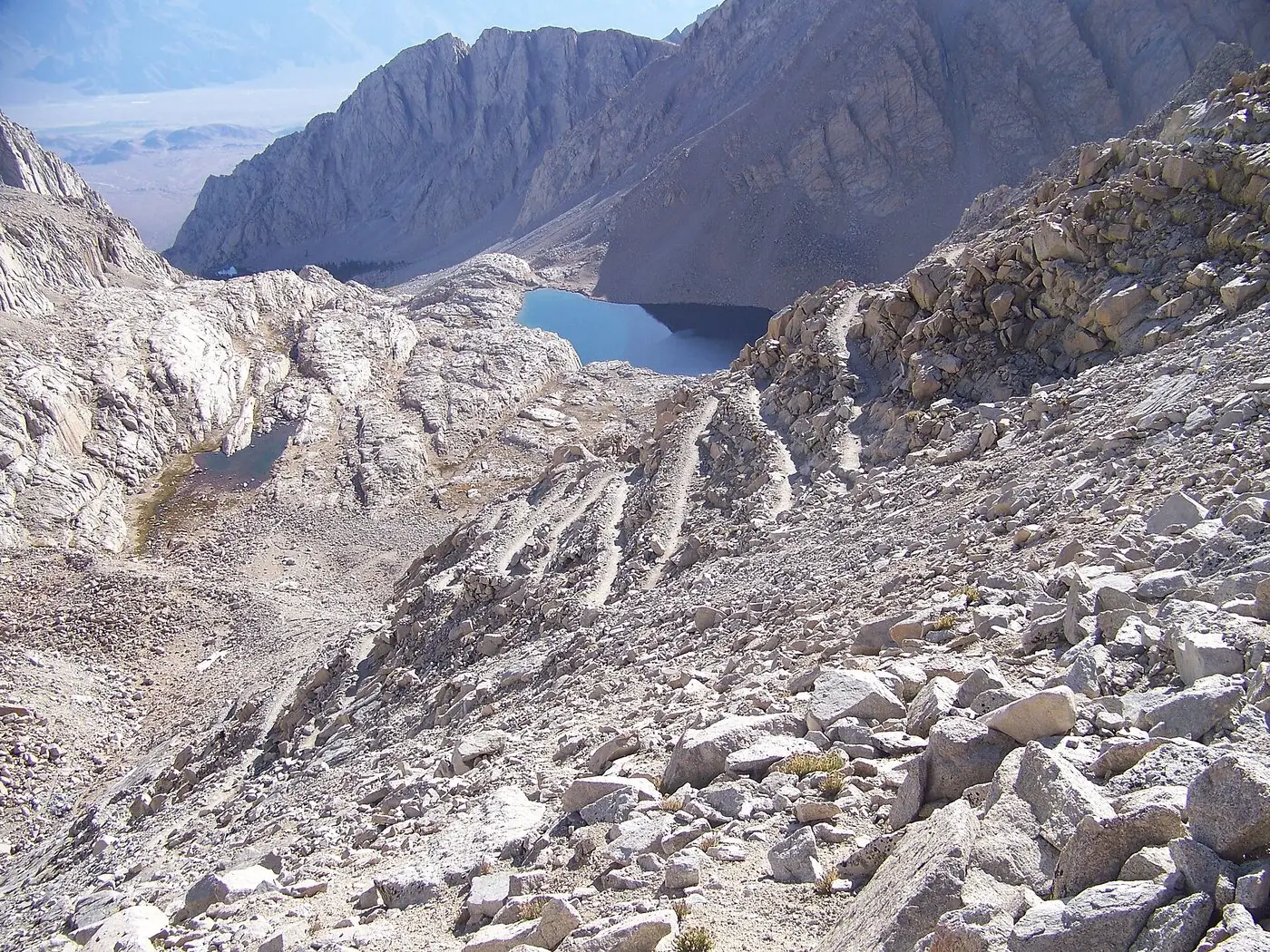 The Mount Whitney Trail switchbacking up a vast slope of pale granite talus in the Sierra Nevada, with Consultation Lake and the Owens Valley haze beyond