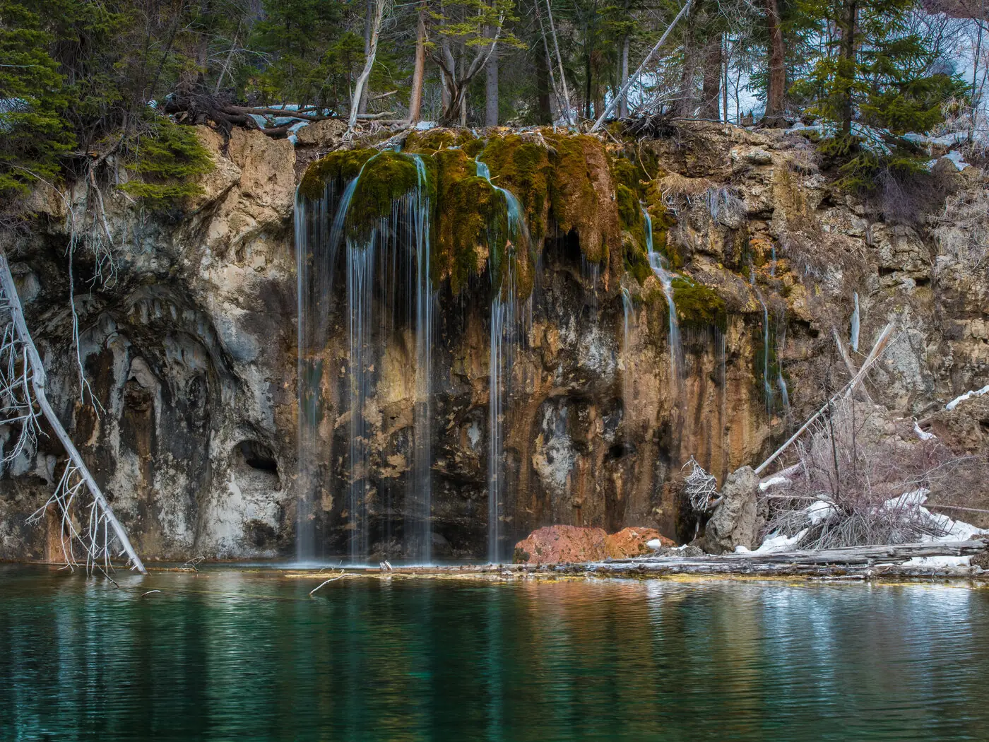 The turquoise water of Hanging Lake with thin waterfalls pouring over a mossy travertine ledge into it, bare trees and patches of snow on the cliff above