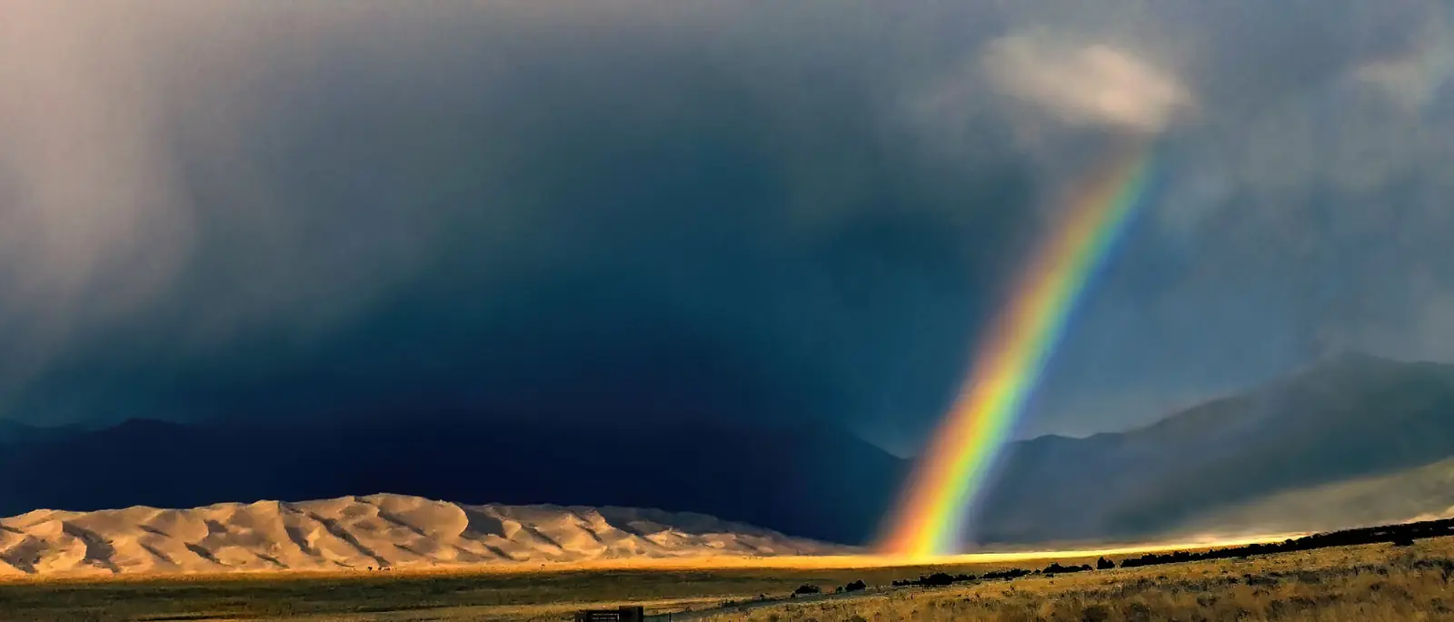 A dark curtain of rain falling from a storm over the Sangre de Cristo Mountains with a bright rainbow at its edge, the Great Sand Dunes lit gold beneath it