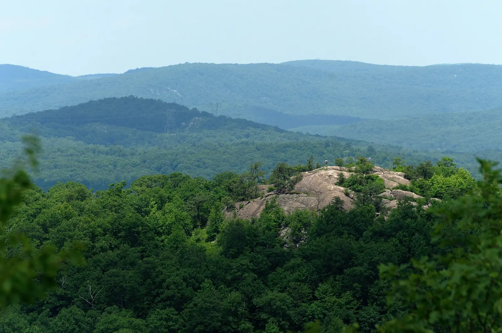 The bare rock dome of Wyanokie High Point rising out of dense green forest, with two tiny figures standing on the summit and wooded ridges behind