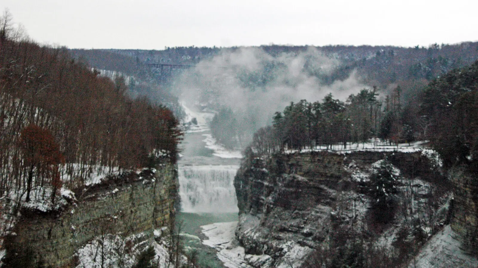The Genesee River gorge at Letchworth State Park in winter, snow on the layered cliff walls with mist rising from the falls and a railway bridge in the distance