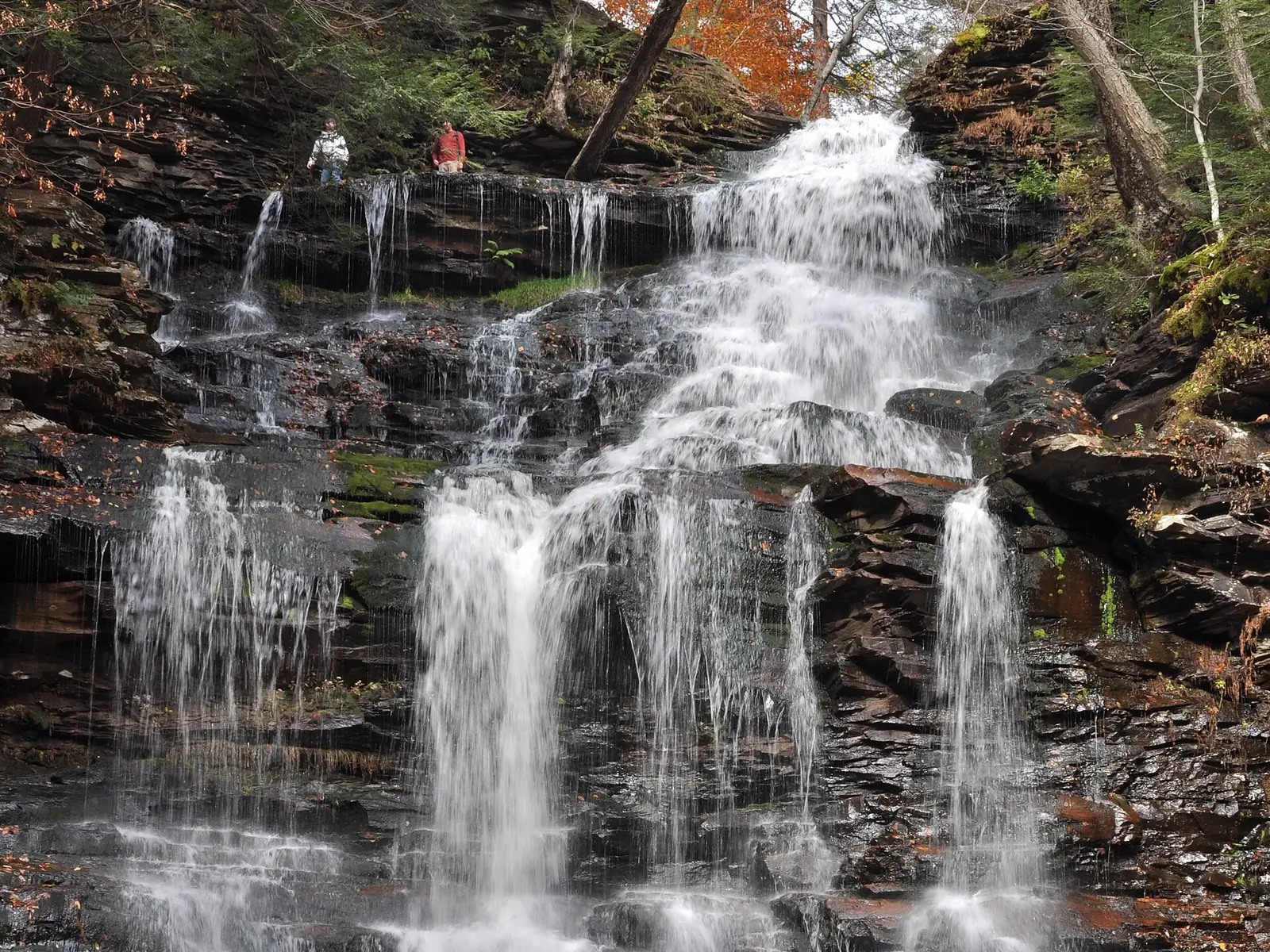 A tall, wide cascade breaking into many white threads down a staircase of dark wet ledges, two people standing at the top edge for scale and orange autumn leaves above