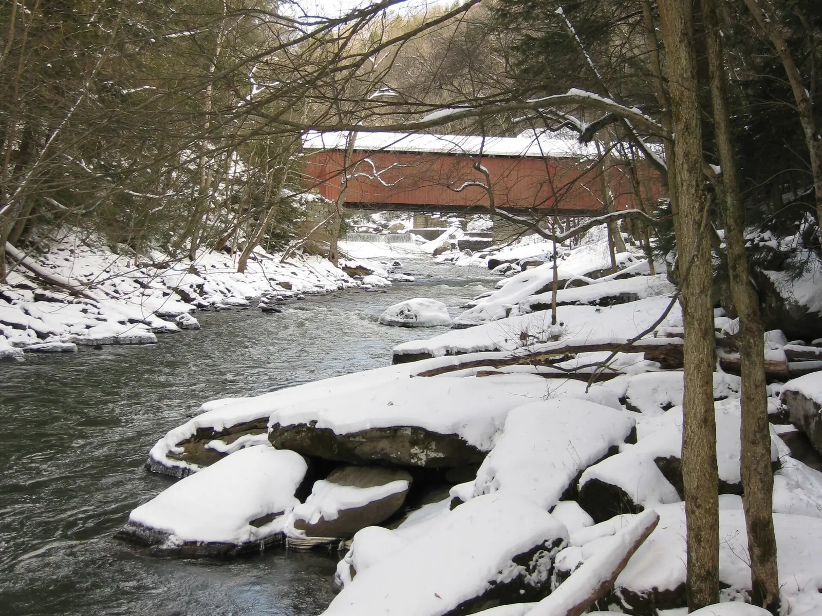 A red covered bridge over a fast creek in winter, with large flat boulders under deep snow filling the near bank and bare trees on both sides