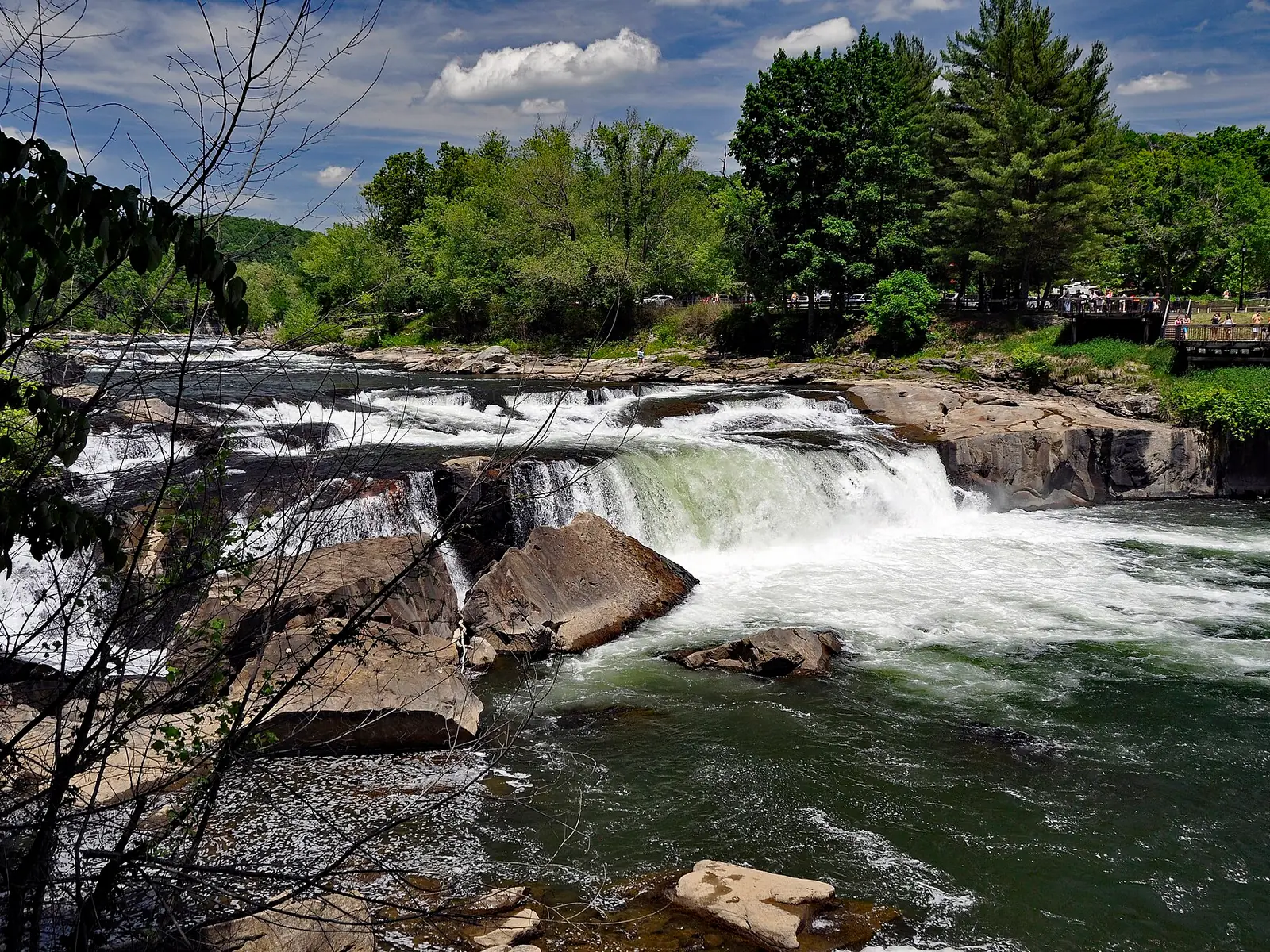 A wide, low waterfall spanning the full width of a river, white water dropping over a ledge of gray sandstone with a wooden viewing deck and pines on the far bank