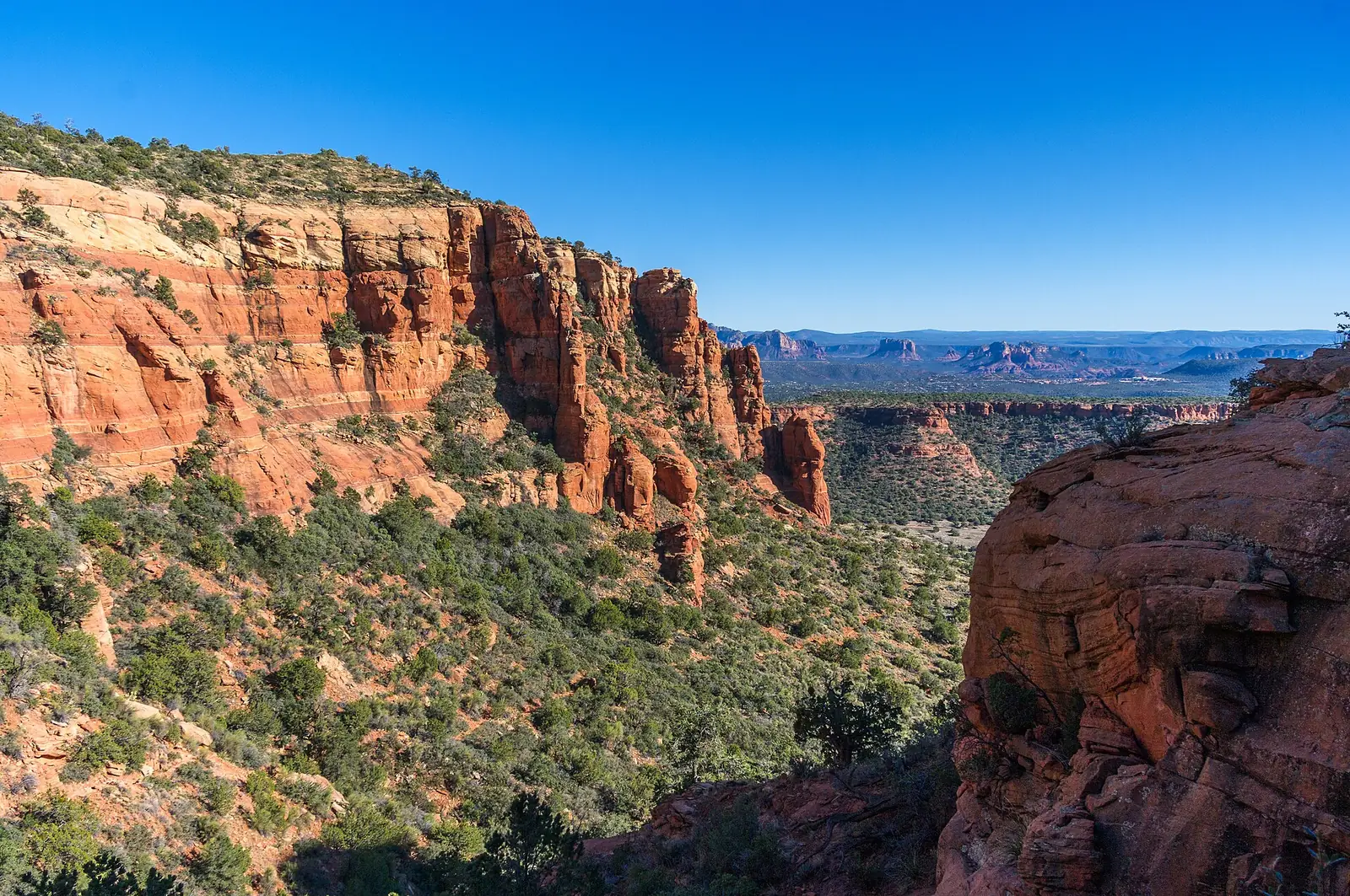 The view from high on Bear Mountain near Sedona, layered red cliff bands falling away to the left and a wide green valley with distant red buttes on the horizon under a clear blue sky