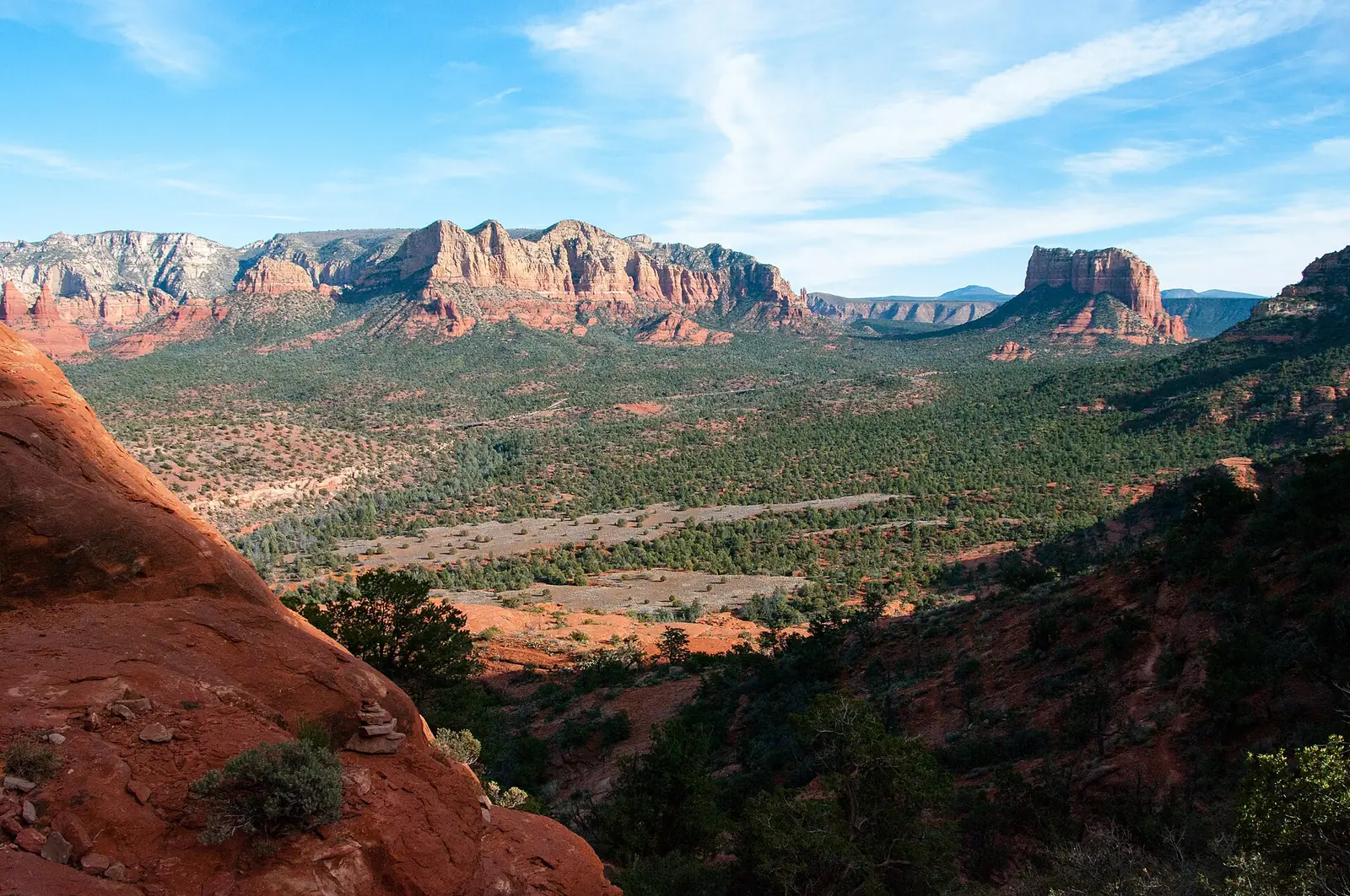The view east from the Cathedral Rock Trail over a wide green valley toward long red cliff walls and the free-standing block of Courthouse Butte, with slickrock and a small cairn in the foreground