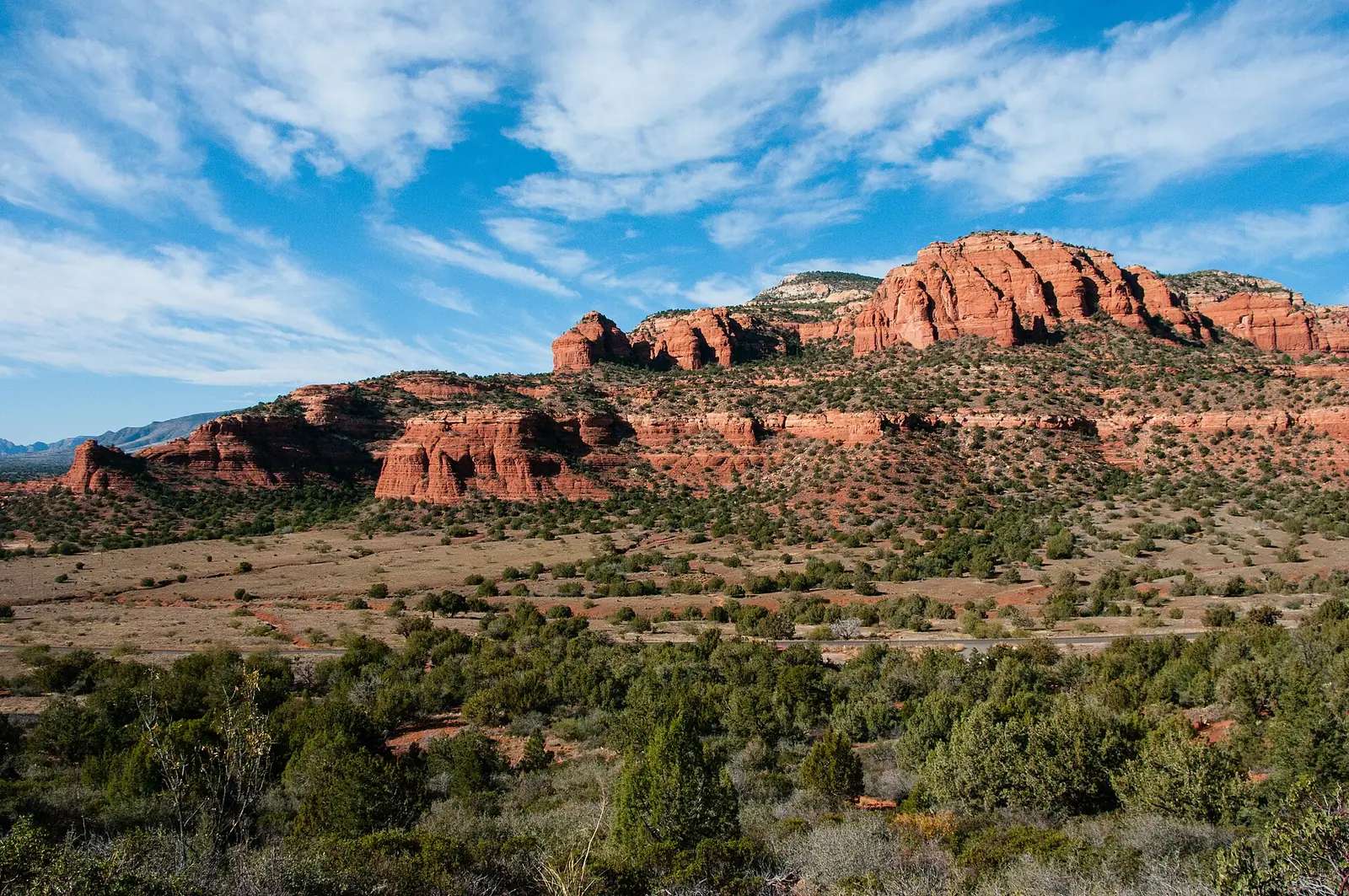 Bear Mountain seen from the Doe Mountain Trail, a broad red sandstone massif of stacked cliff bands rising above a plain of juniper and scrub, with a paved road crossing the middle distance under a blue sky with high cloud