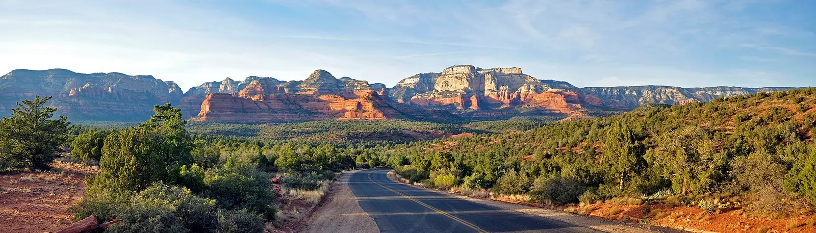 A panorama of the red rocks near Sedona seen from a quiet paved road, a long line of red sandstone buttes and cliffs capped with pale cream rock rising above a broad plain of green juniper