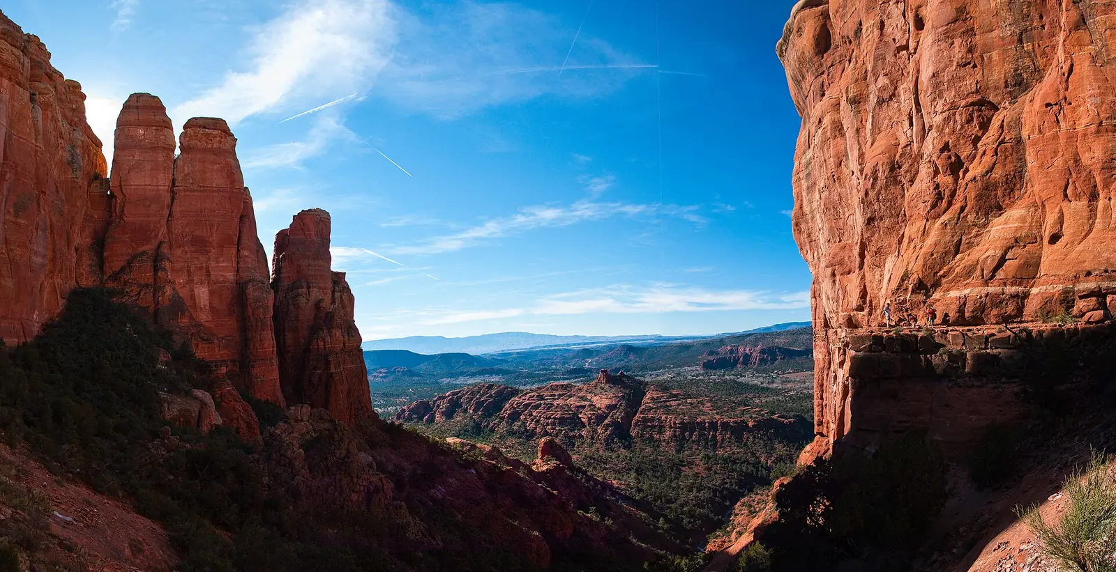 The saddle on the Cathedral Rock Trail in Sedona, red sandstone spires rising on the left and a sheer wall on the right with hikers standing on a ledge, looking out over green valley and distant mesas