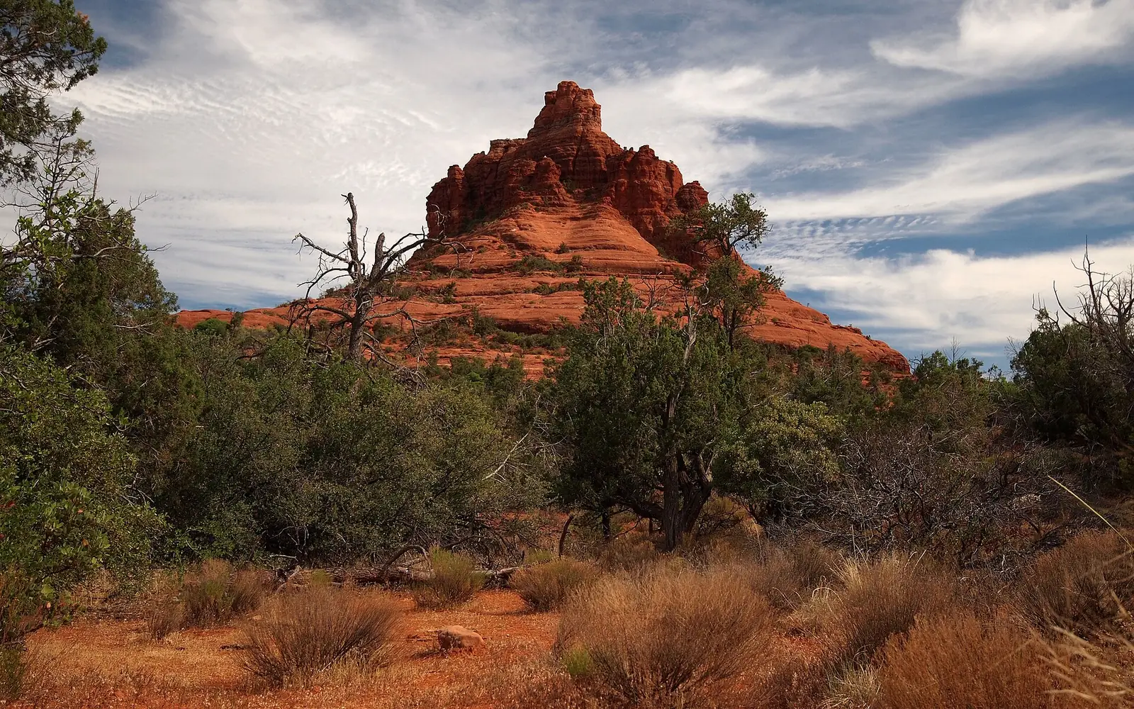 Bell Rock near Sedona, a bell-shaped red sandstone butte rising above juniper scrub and dry grass under a streaked blue sky