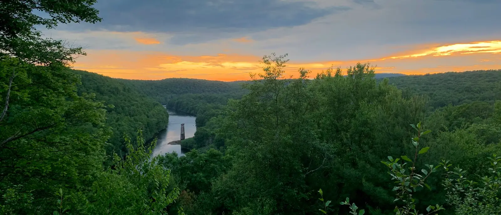 Sunset over the Lehigh Gorge seen through a gap in summer foliage, the river winding between forested ridges with an old stone bridge pier standing in the water