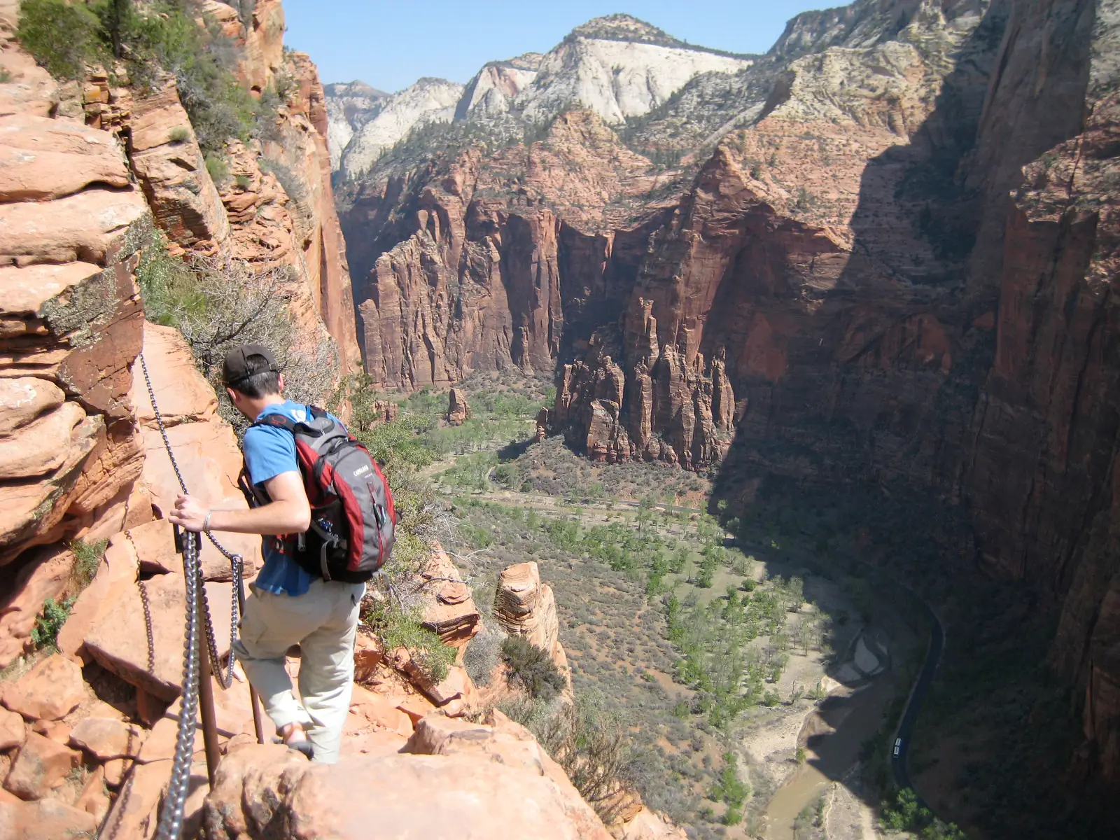 A hiker holding a chain bolted to the rock on the Angels Landing spine, looking down more than a thousand feet into Zion Canyon