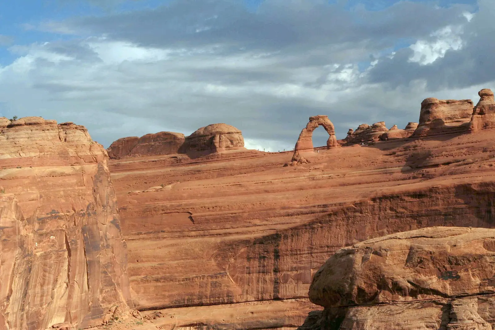 Delicate Arch standing on the rim of a slickrock bowl with tiny figures of hikers nearby, under a bank of cloud
