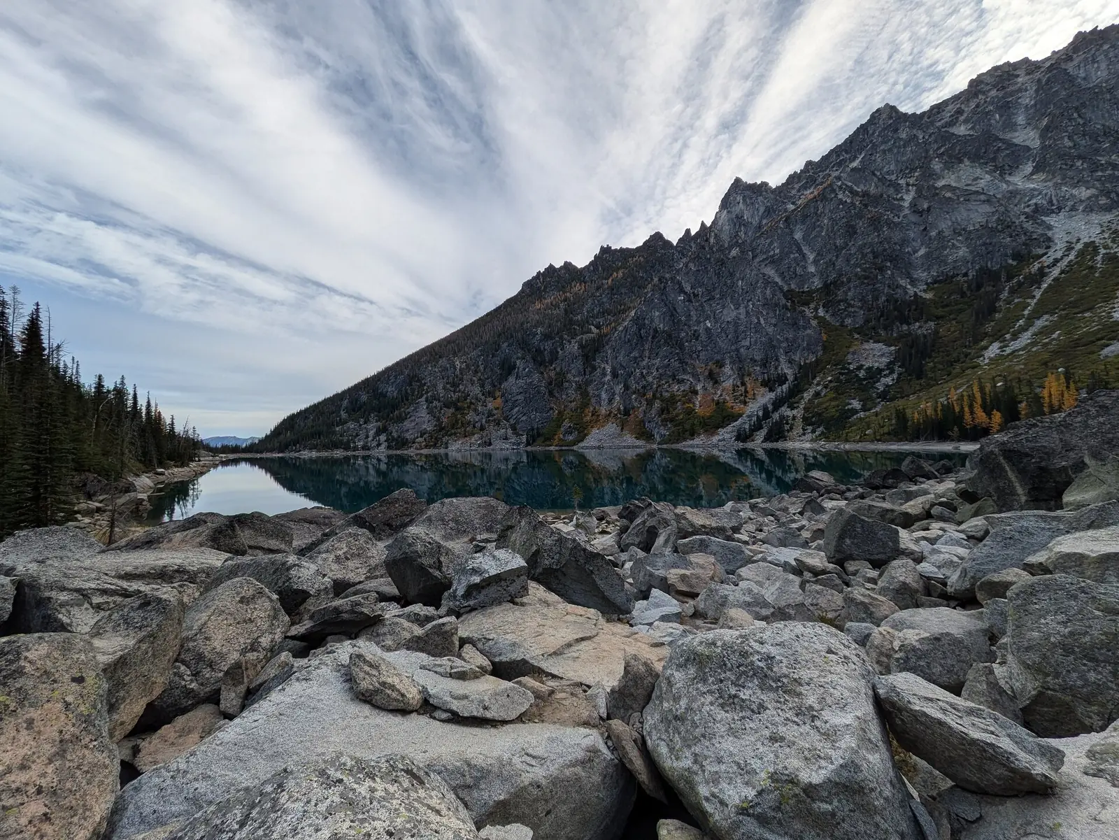 A still turquoise alpine lake below a jagged gray granite peak, with a boulder field in the foreground and gold larches scattered across the far slope